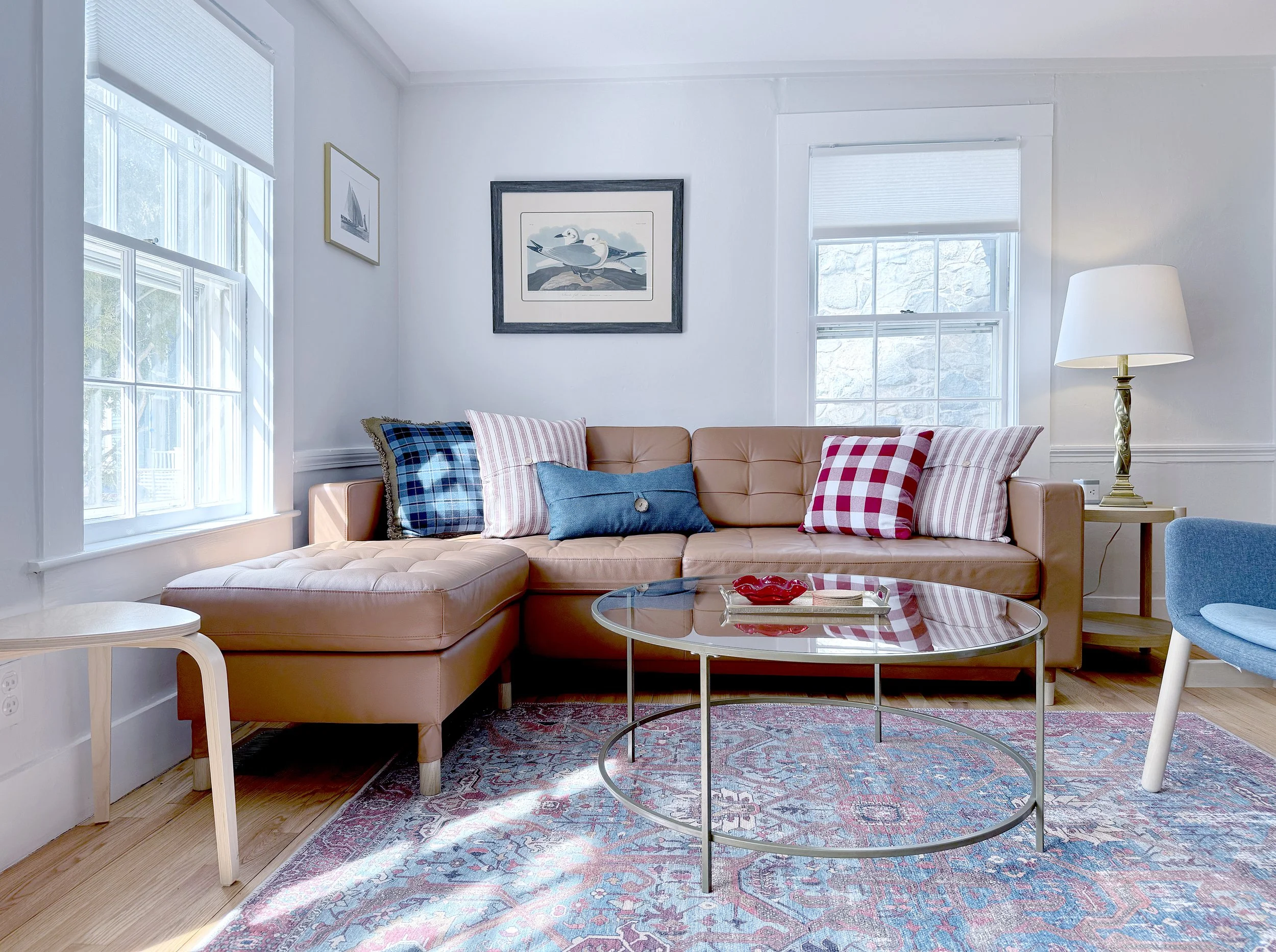 Living room with a tan sectional sofa, patterned cushions, a glass coffee table, and a floor lamp. Two windows let in natural light, and artwork decorates the white walls.