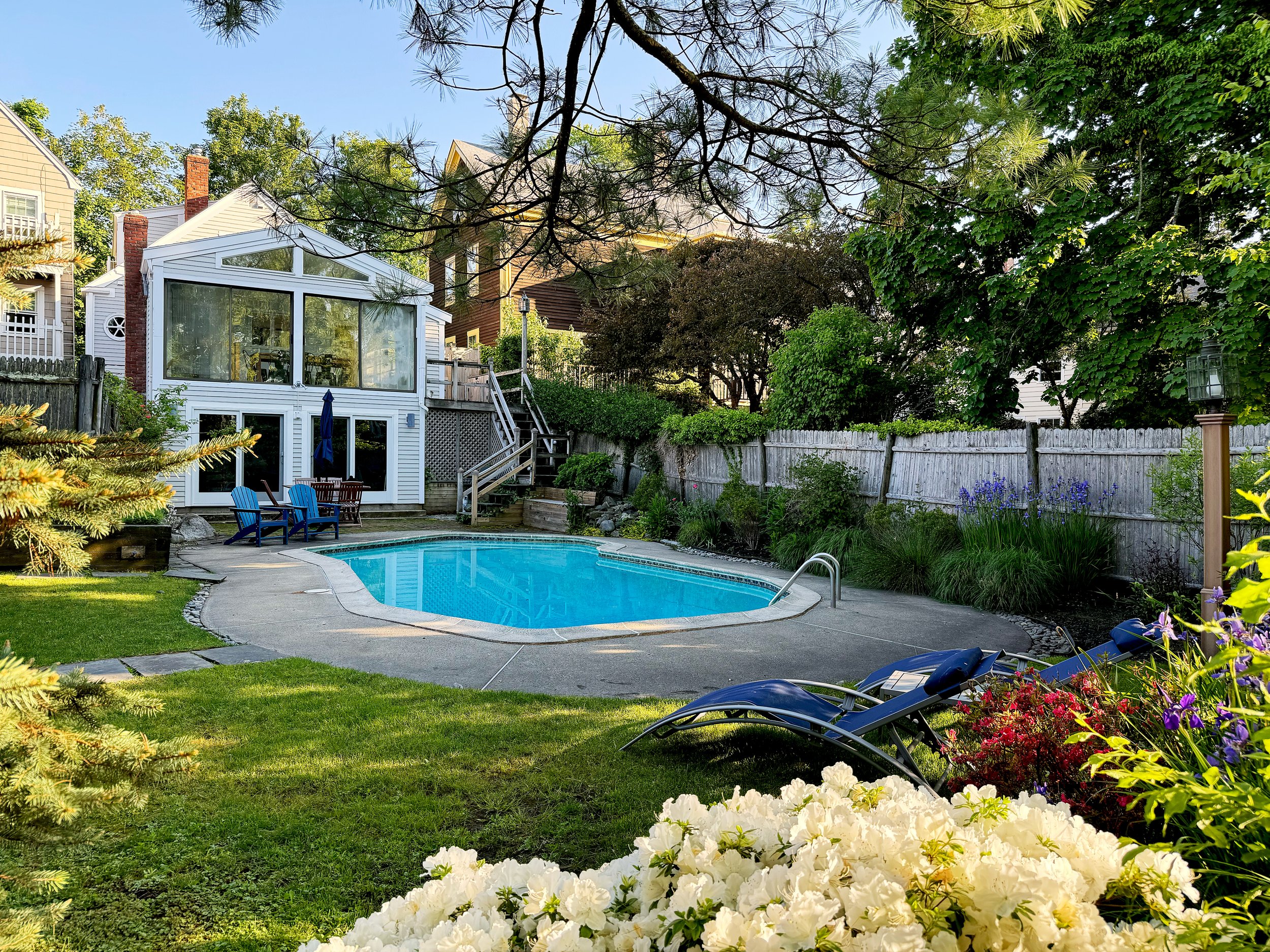 Backyard with a swimming pool, surrounded by a garden with trees and colorful flowers, a fenced yard, and a house with a screened porch and stairs.