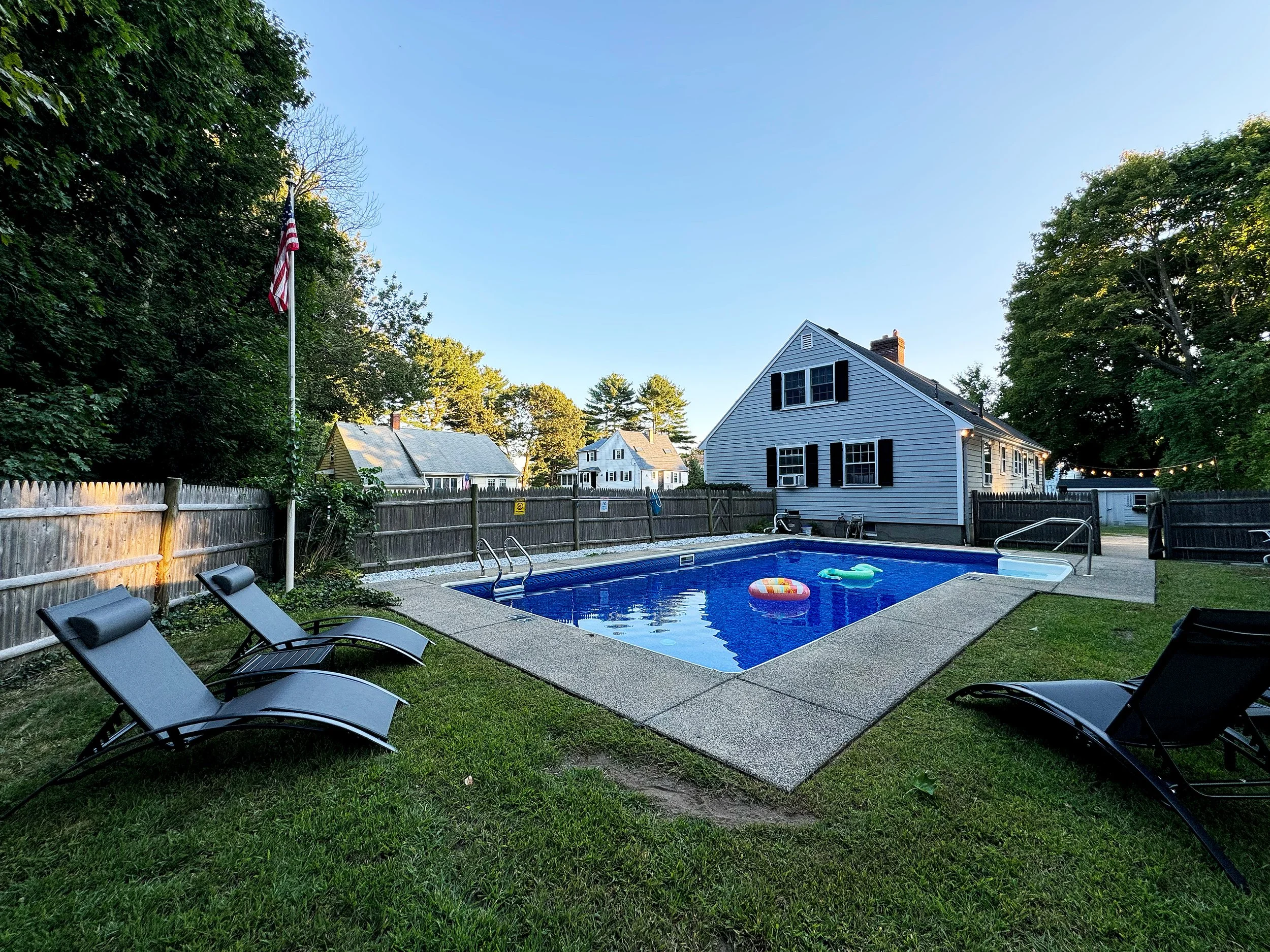 Residential backyard with a swimming pool, four lounge chairs, a flagpole, a house, and trees.