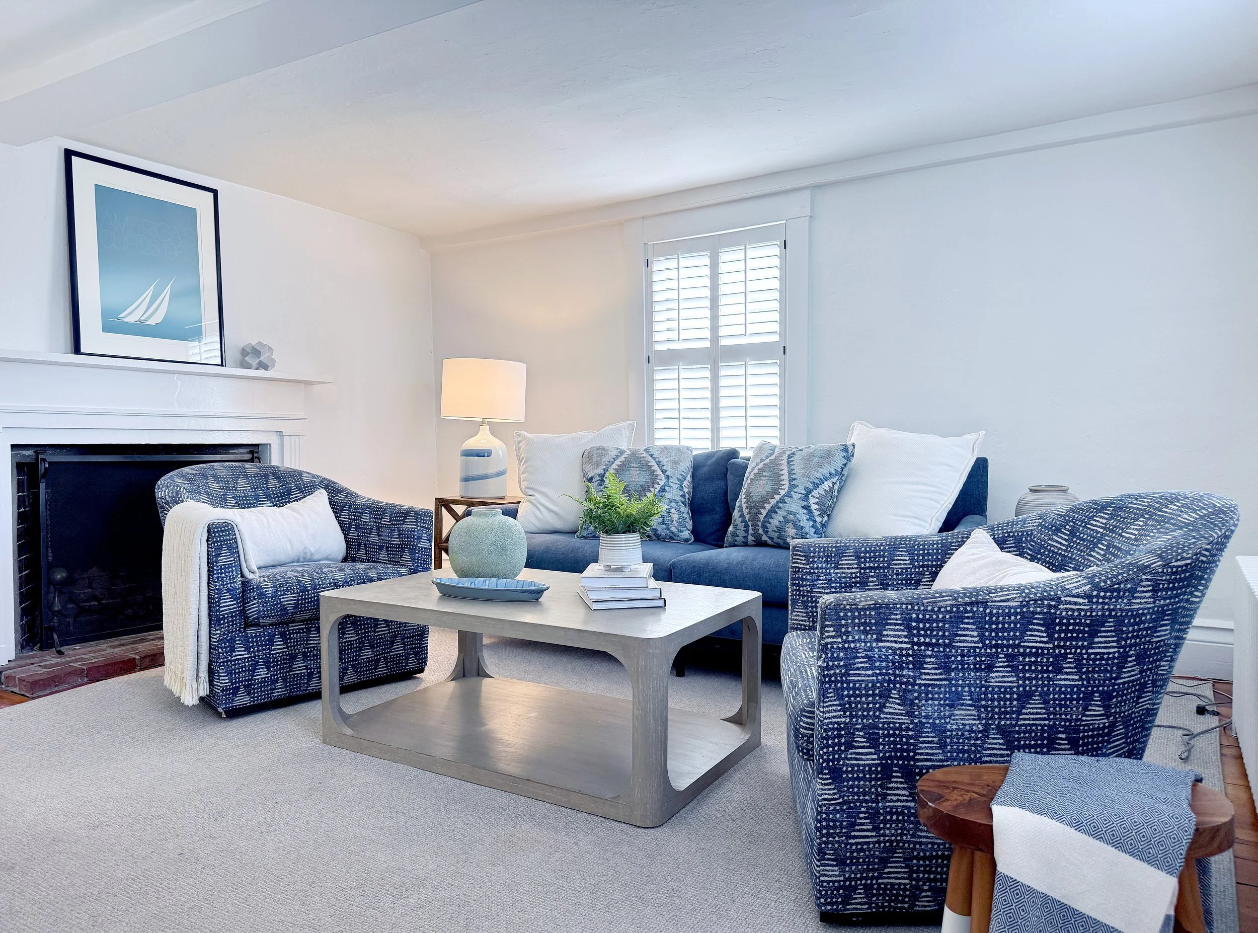 A living room with a white fireplace, blue patterned armchairs, a matching sofa with pillows, a coffee table with decorative items, a table lamp, a window with shutters, and neutral-colored walls and carpet.