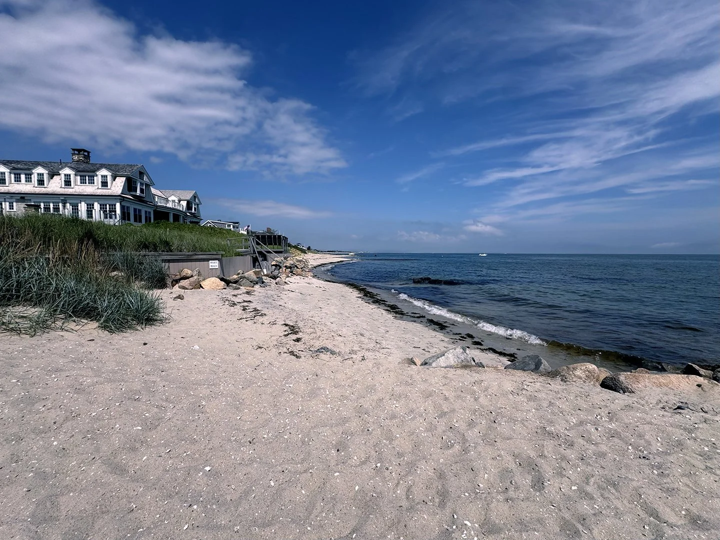 Sunny beach with houses on a grassy dune, sandy shoreline, rocks, ocean waves, and partly cloudy blue sky with streaks of white clouds.