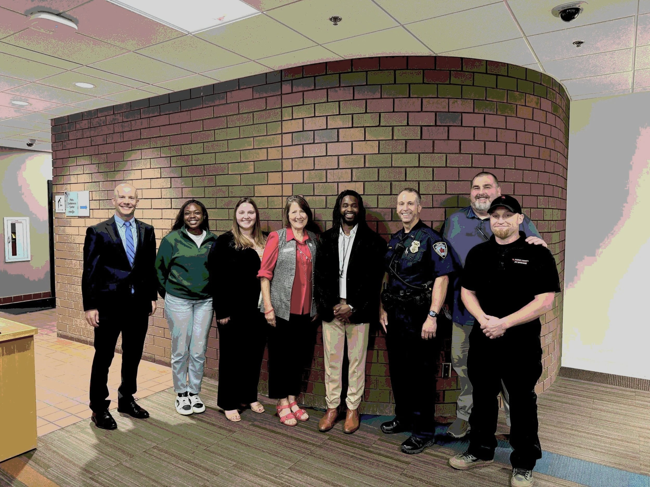 Group of nine diverse people, including a man in a police uniform and others in casual or business attire, standing together indoors in front of a curved brick wall and smiling.