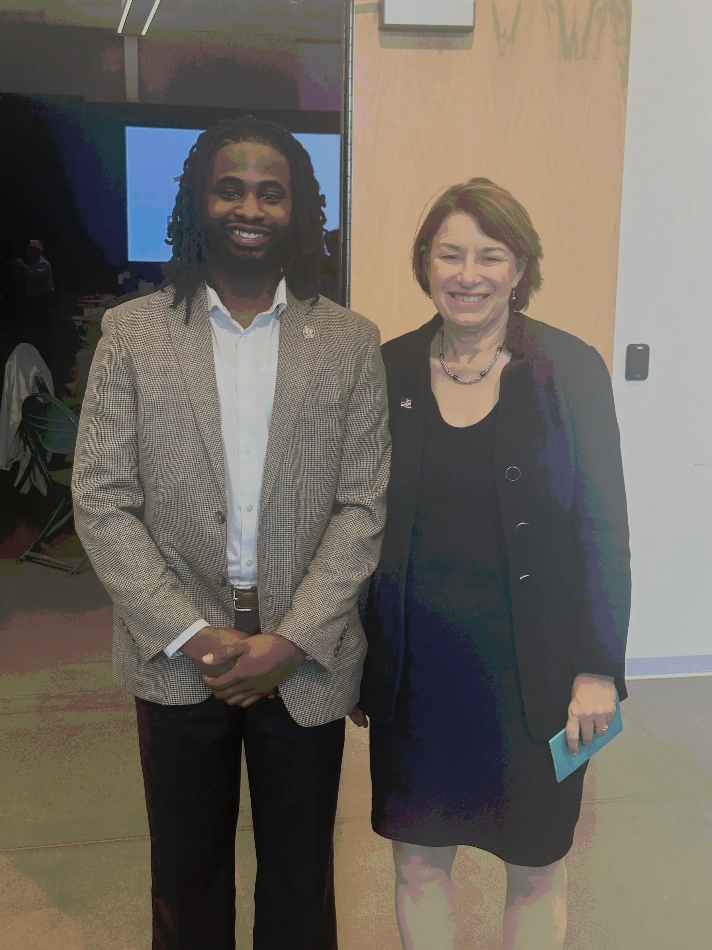 Two women standing together, smiling for the photo. The woman on the left has dark skin and dreadlocks, wearing a beige blazer over a white shirt. The woman on the right has light skin and short hair, wearing a black dress with a black blazer and holding a small light blue object in her right hand.