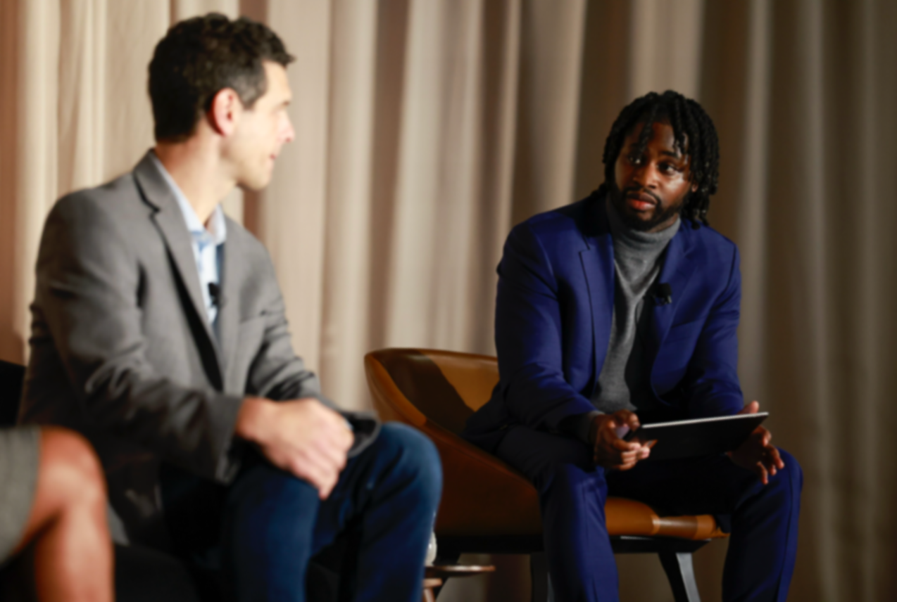 Two men in suits sitting and having a conversation in a room with beige curtains.