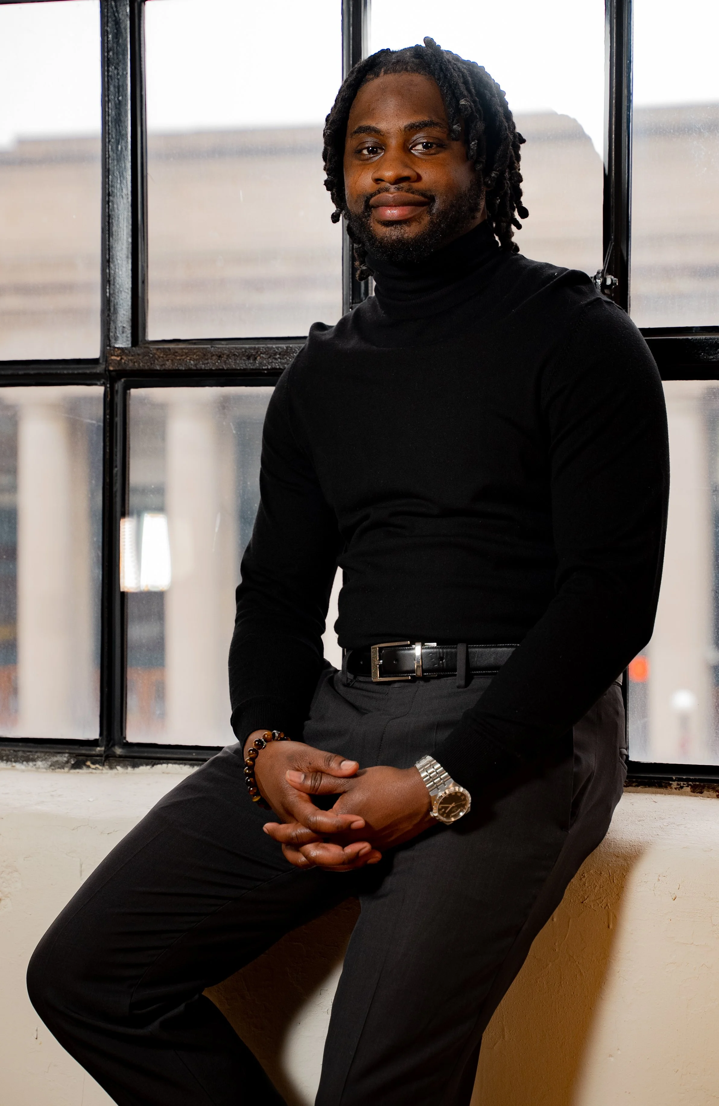 A man with dreadlocks wearing a black turtleneck, gray trousers, and a wristwatch, sitting on a windowsill in front of a large industrial-style window.