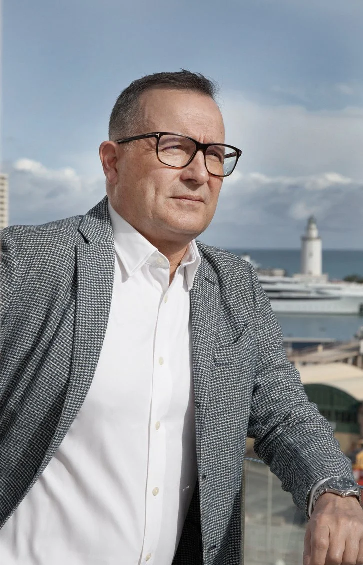 A man wearing glasses and a checked blazer stands outdoors with a city and harbor in the background, looking to the side.