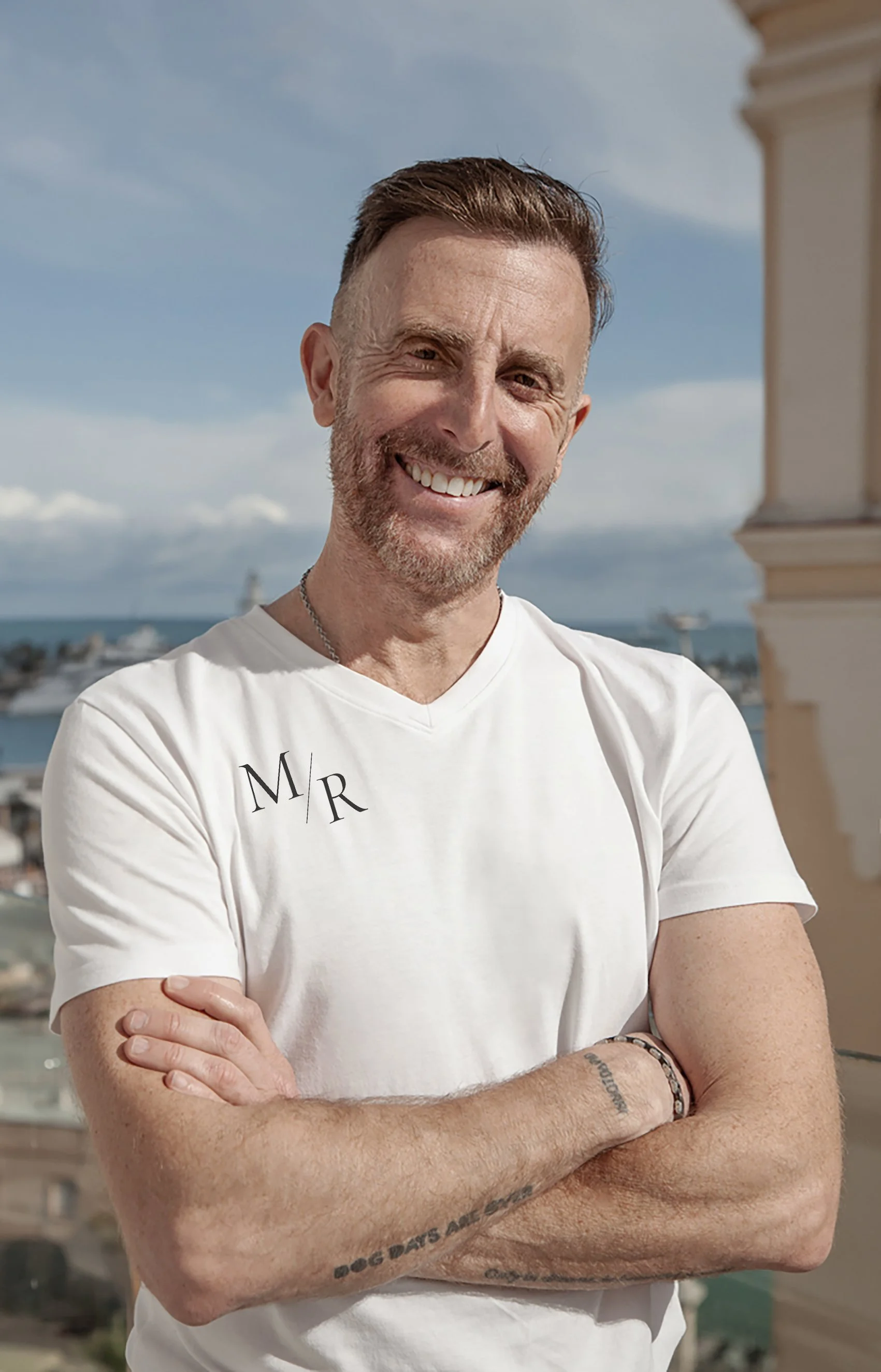 Man with short brown hair and a beard smiling outdoors, wearing a white T-shirt with the initials 'M R,' standing against a cityscape background with a body of water and a cloudy sky.