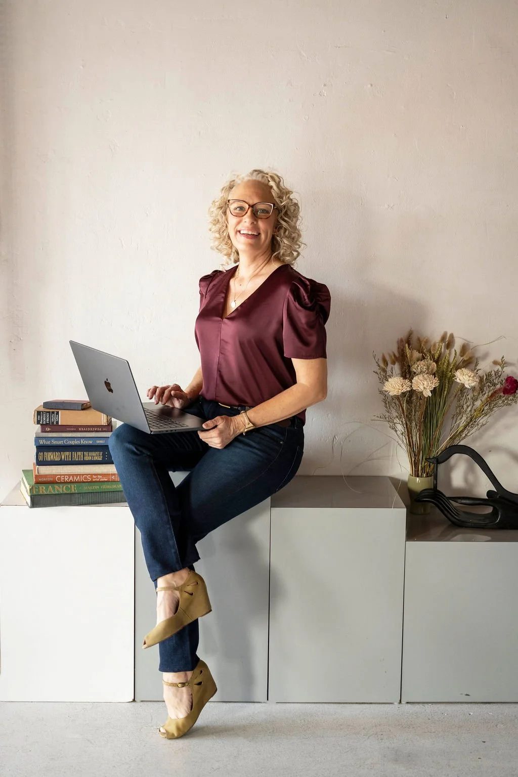 A smiling woman with curly blonde hair, glasses, and wearing a maroon satin blouse and dark jeans, sitting on a white cabinet with a laptop in her lap in a modern, minimalist room with a white wall, a stack of books, and a vase of dried flowers.