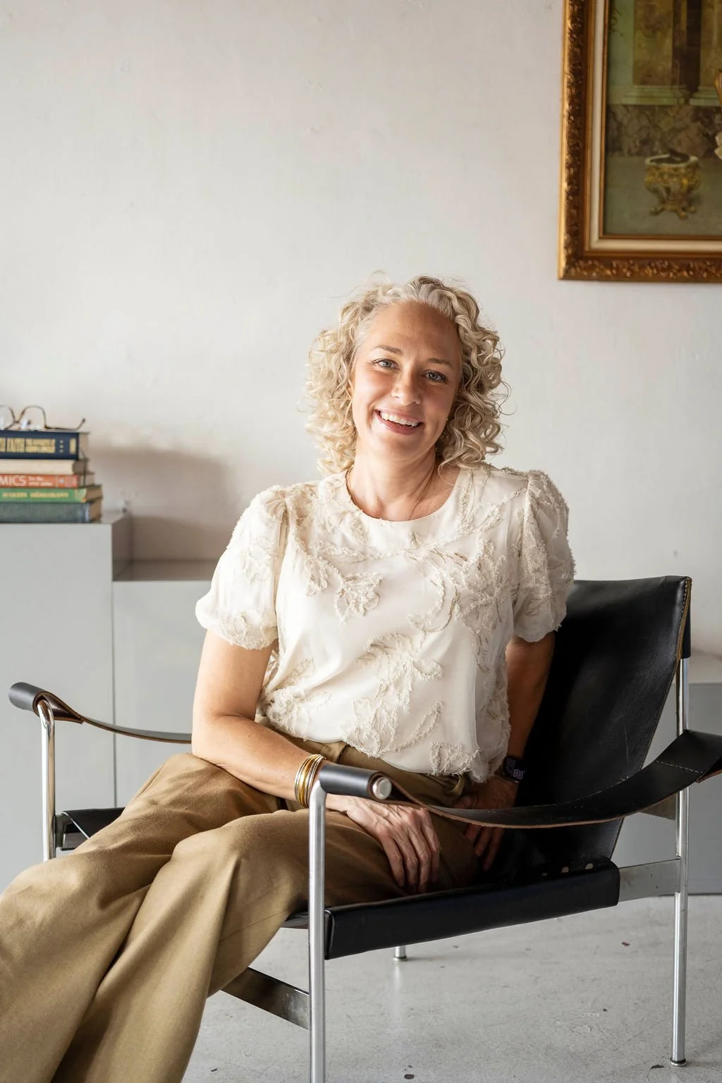 A smiling woman with curly blonde hair, wearing a cream-colored embroidered blouse and gold pants, sitting on a black leather and metal chair in a modern room with white walls, a wall painting, and a white cabinet with stacked books.