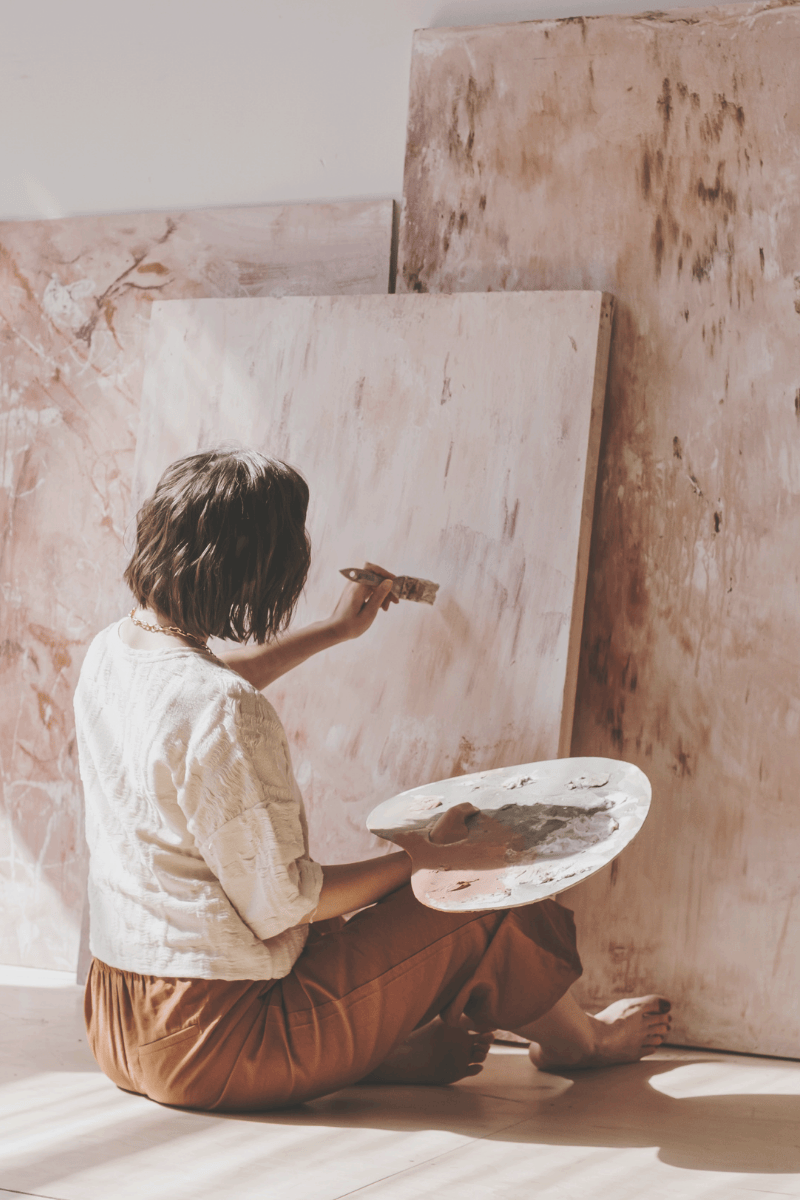 Woman sitting on the floor painting on a large canvas, holding a palette with paint in her lap and a brush in her hand, with other large canvases leaning against the wall.
