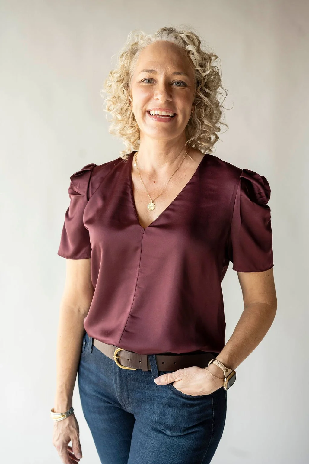 A woman with blonde curly hair smiling, wearing a maroon short-sleeve satin blouse, blue jeans, and accessorized with jewelry, standing against a plain light background.