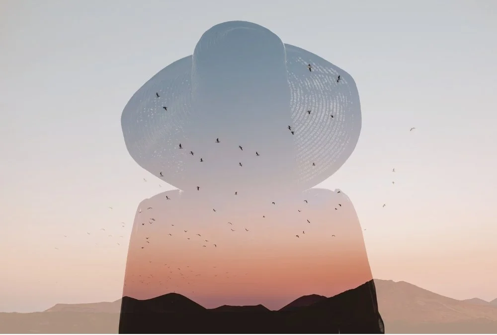 A man wearing a wide-brimmed hat is seen in a double exposure photograph with mountains and a flock of birds at sunset.
