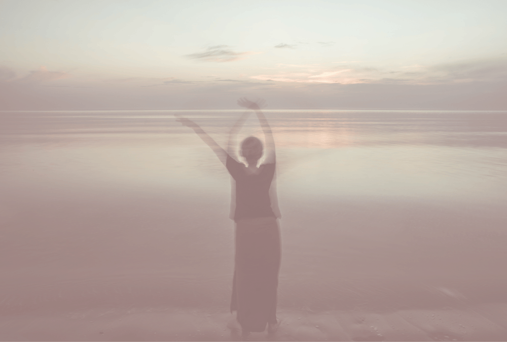A silhouette of a person standing on the beach with arms raised, facing the ocean during sunset, with the image having a blurry, dreamy effect.