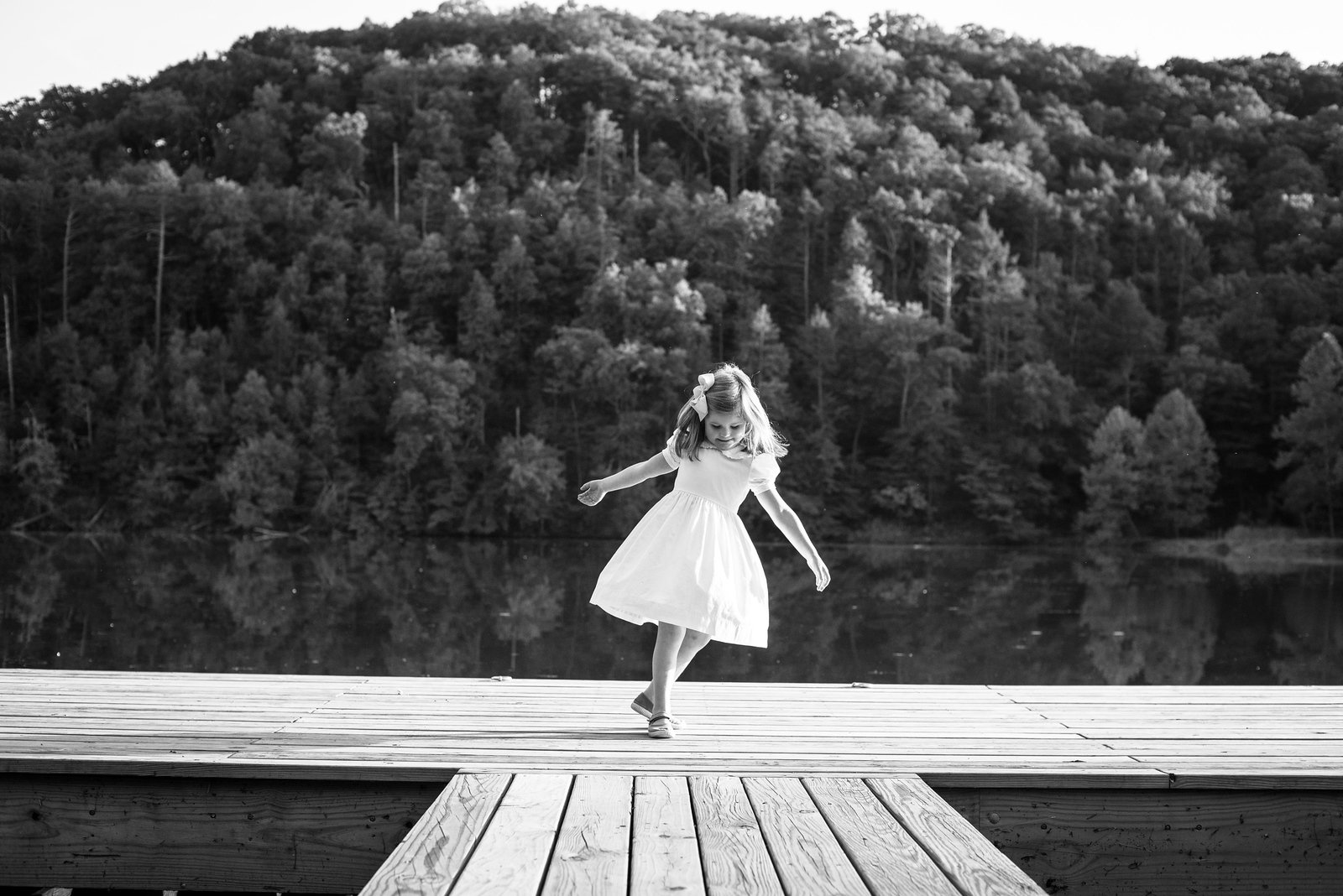 Young girl in white dress twirls on wooden dock by lake with forested hills behind, black and white