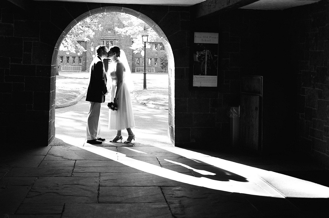 Bride in white gown and veil kisses groom in suit under stone archway with sunlight shadows