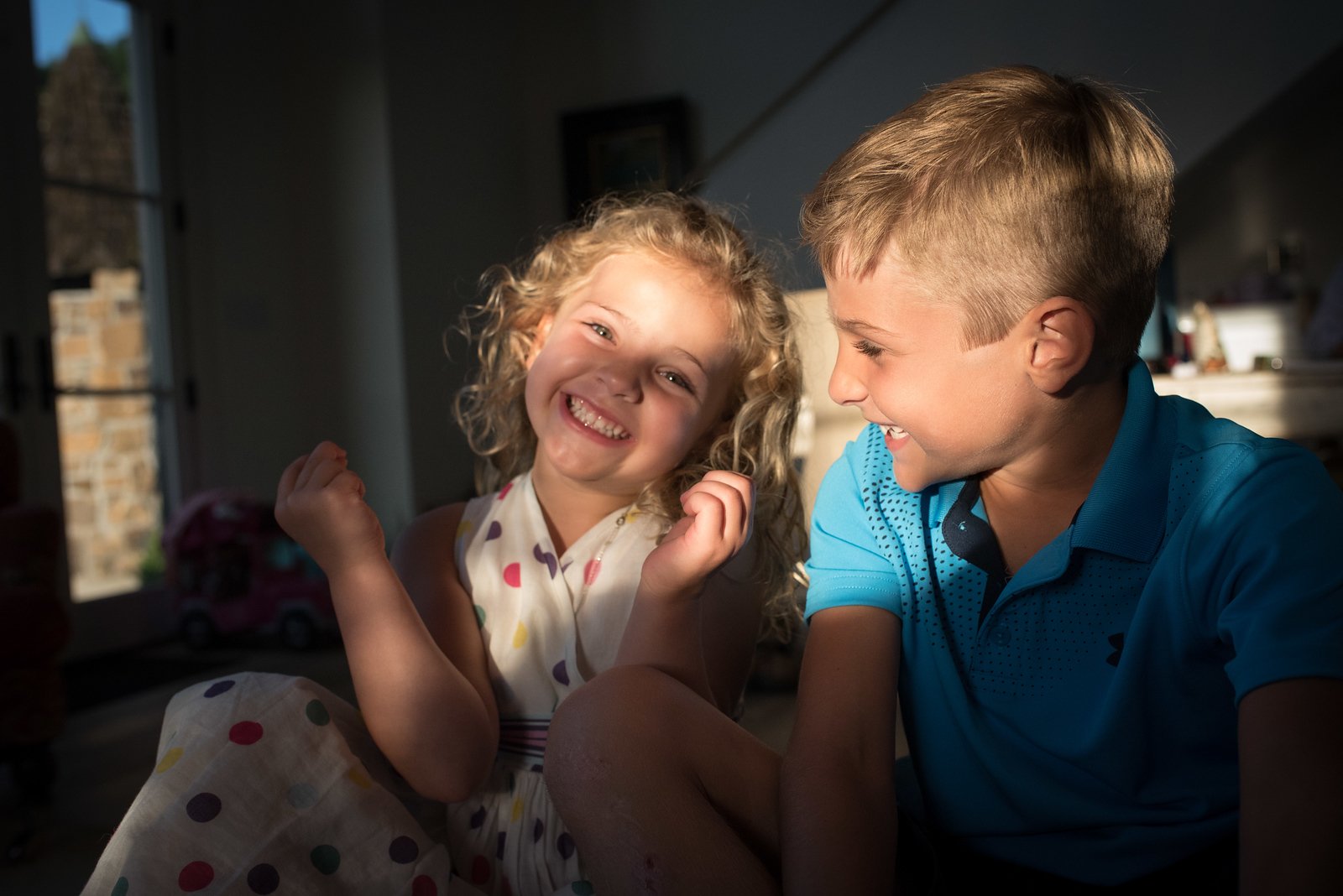 Smiling curly-haired girl in polka-dot dress and boy in blue polo shirt, sunlit room.