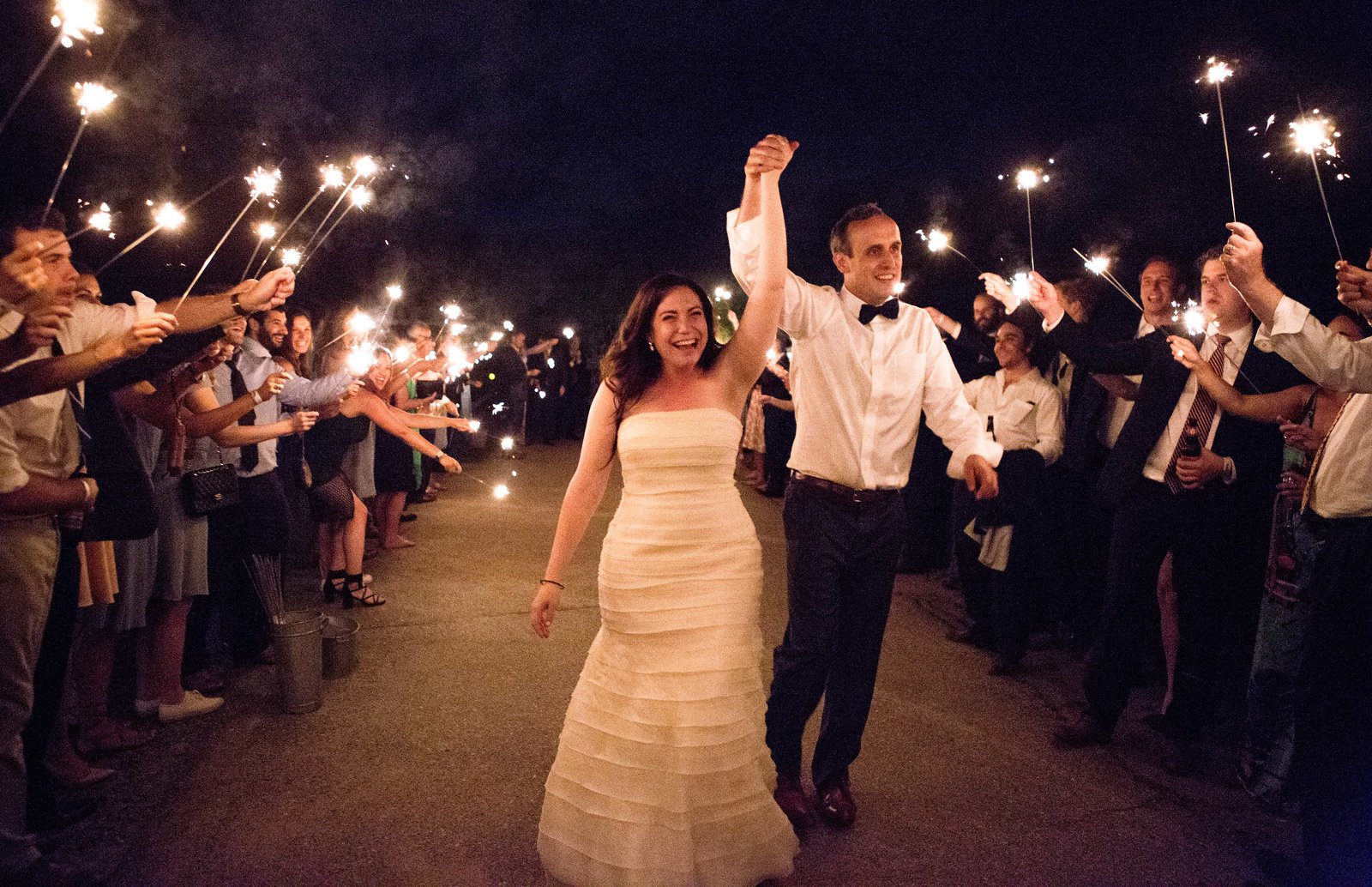 Bride and groom walk hand-in-hand through sparkler tunnel held by wedding guests at night