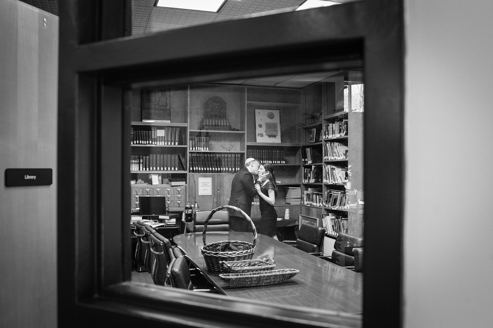 Black-and-white photo of couple kissing in library with bookshelves and desk, framed by window.