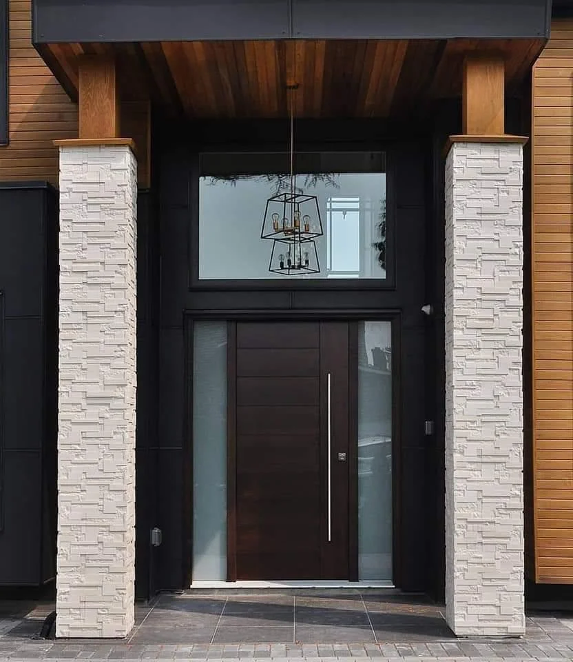 Modern front door with large vertical handle, framed by white stone columns, a large window above, and a wooden ceiling with an artistic chandelier.