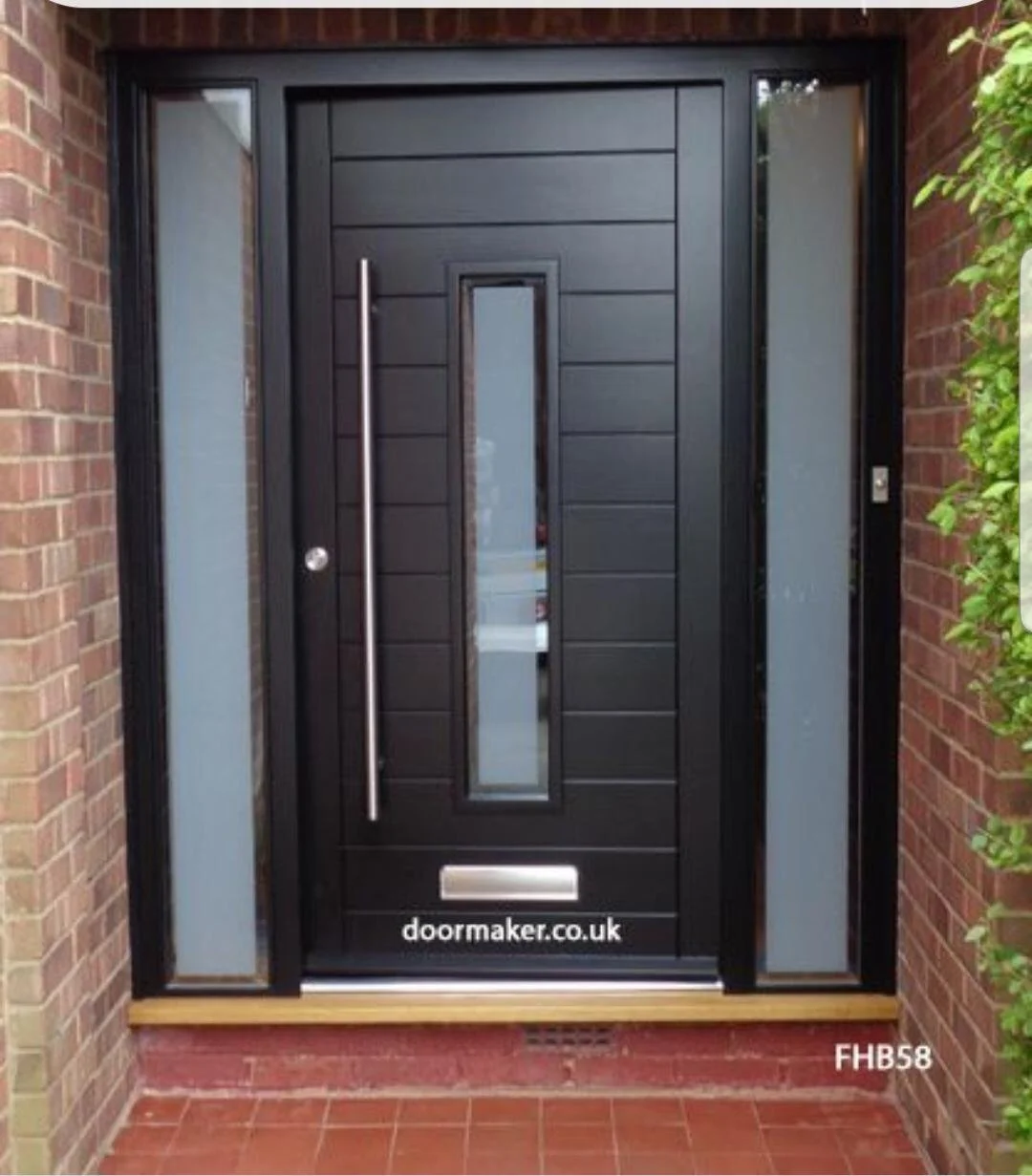 Black front door with vertical window in the center, silver handle, mail slot, and glass sidelight on the right, surrounded by brick walls and green foliage, with a wooden threshold and tiled porch.