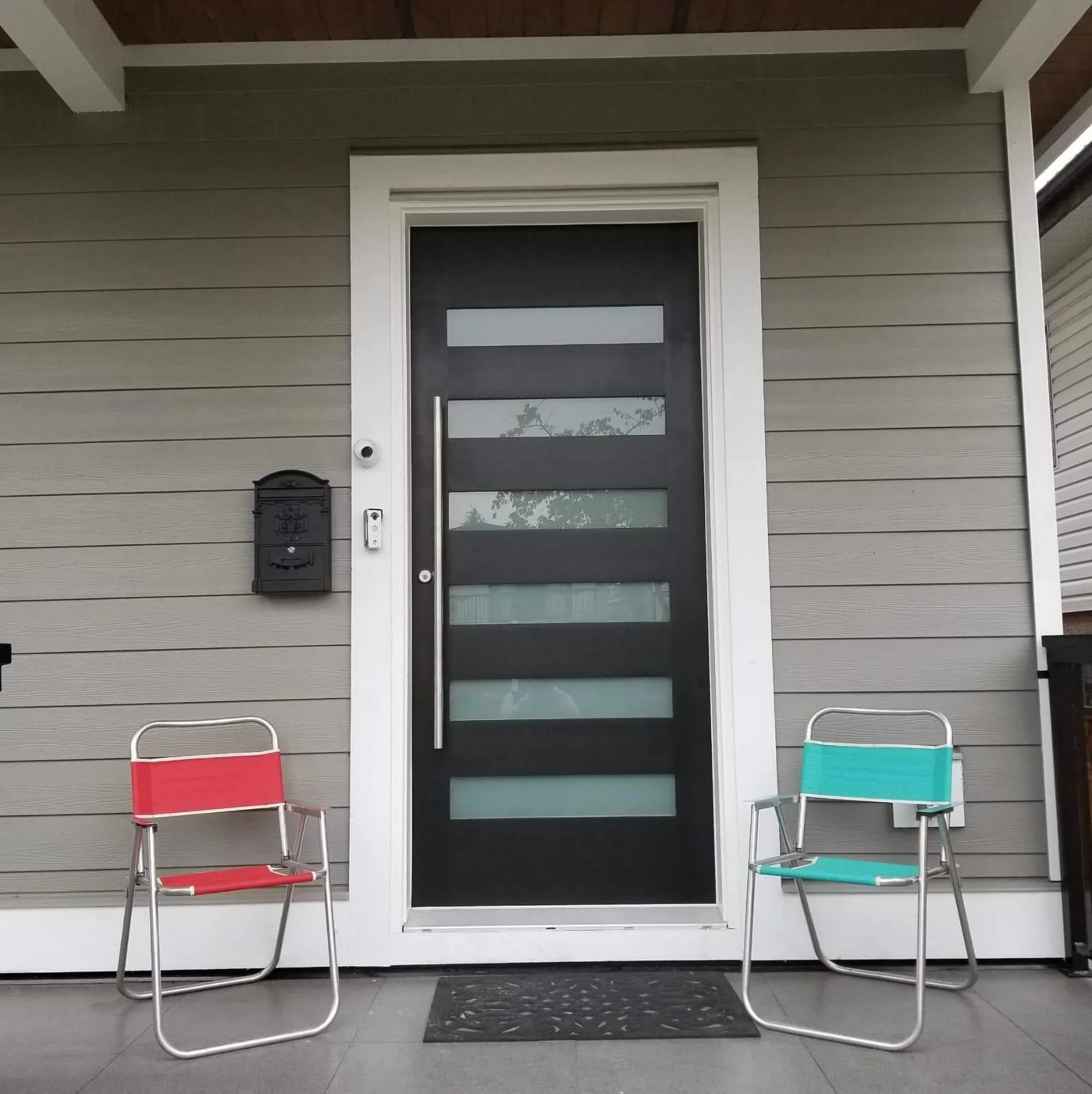 Front porch of a house with a black door featuring five horizontal frosted glass panels and a long vertical handle, flanked by two metal chairs, one with red fabric and the other with teal fabric, and a black mailbox on the wall to the left of the do