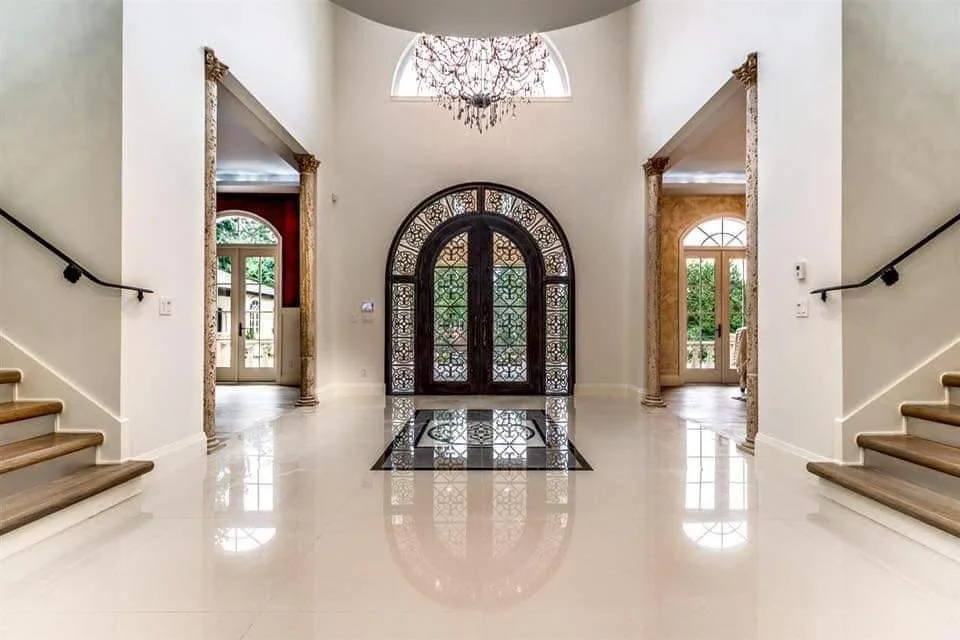 Elegant foyer with white polished tile floors, grand arched dark wooden double doors with intricate glass panels, marble columns framing doorways, and large windows providing natural light.
