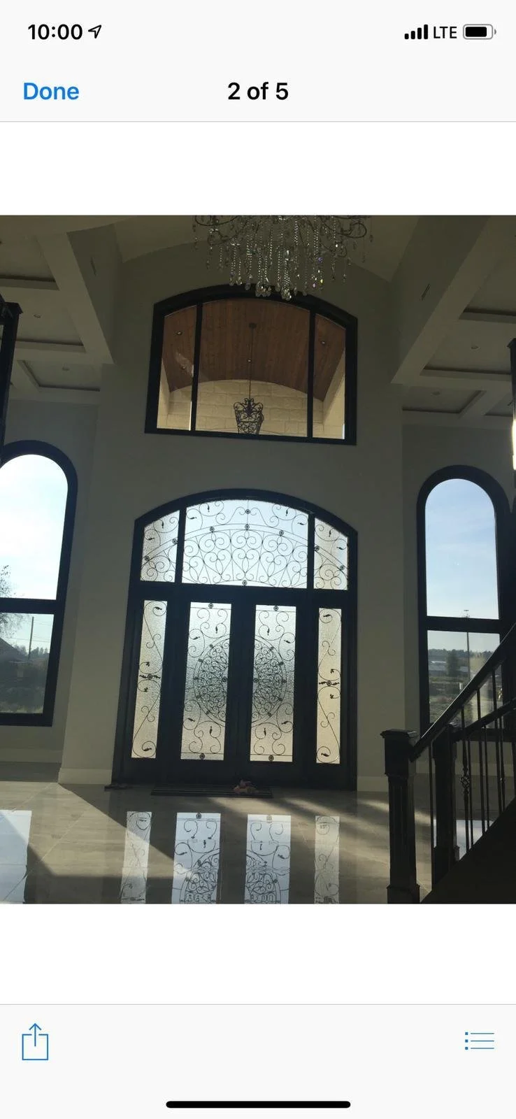 Interior view of a grand entryway with large arched glass door and sidelights featuring intricate wrought iron designs, polished tile floor reflecting sunlight, and a high ceiling with a chandelier and a balcony with a decorative railing.