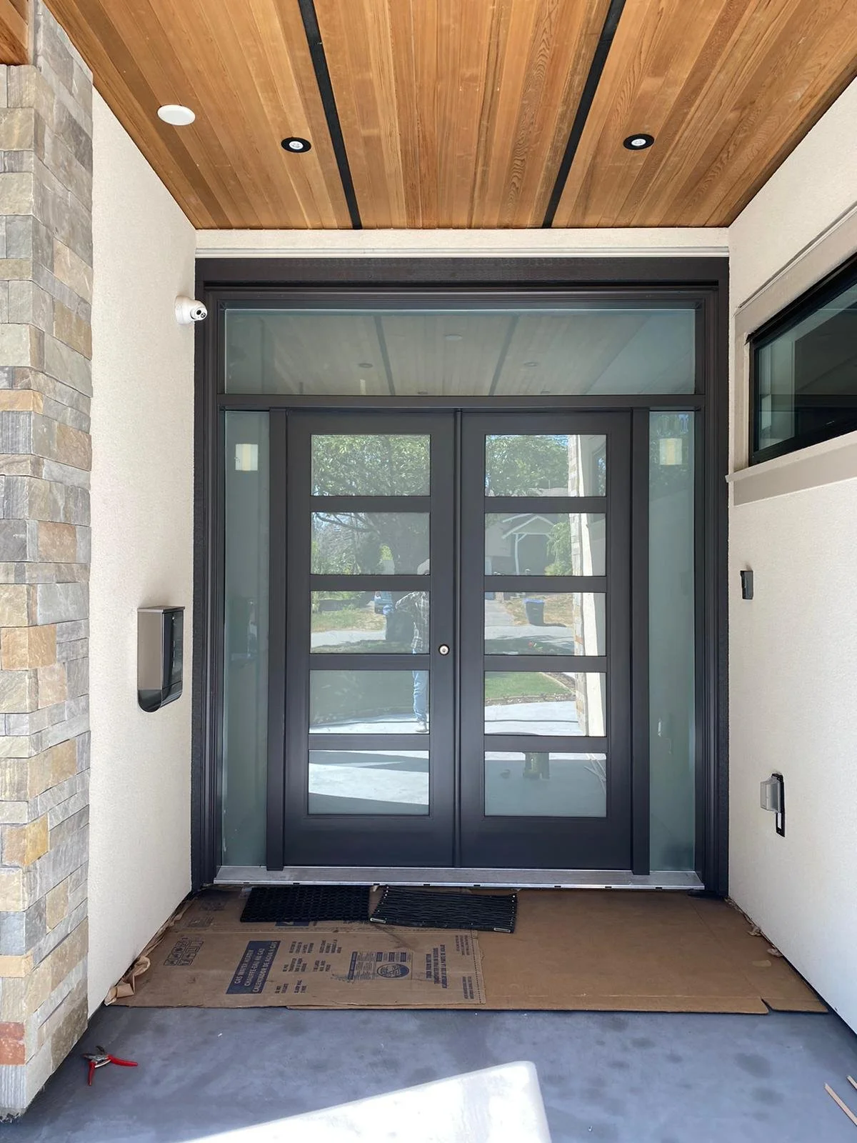 Newly installed front entry door with glass panels, flanked by side windows, with a wooden ceiling above and a stone wall on the left.