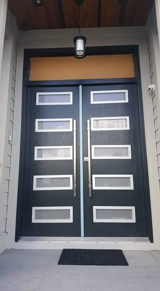 Front view of a modern black double door with horizontal white rectangular accents, metal handles, in a house entrance with a small black doormat, a ceiling light, and beige siding.