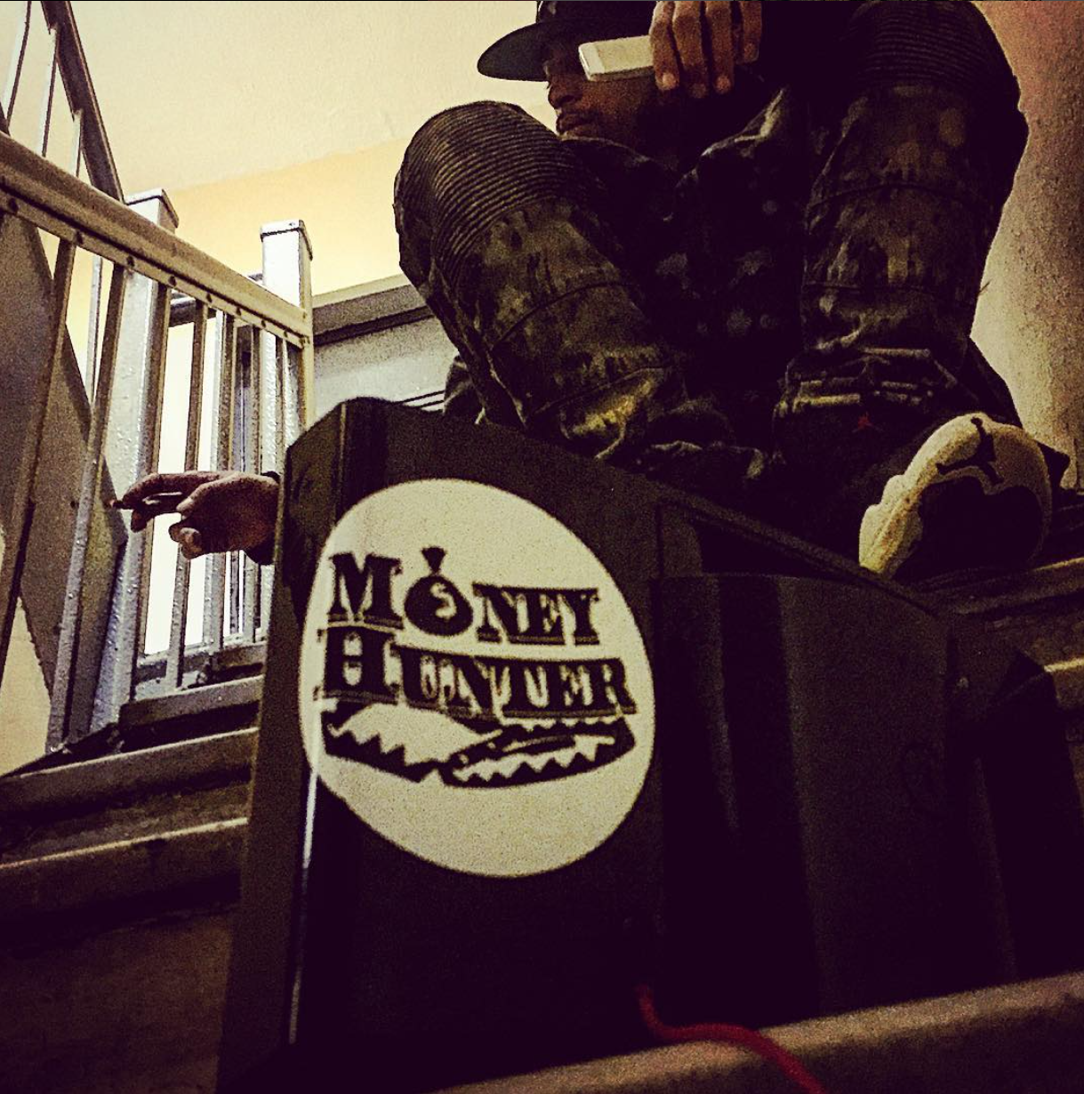 Person sitting on stairs wearing a black hat, camouflage jacket, and sneakers, holding a phone to their ear, with a Monopoly game box in the foreground.