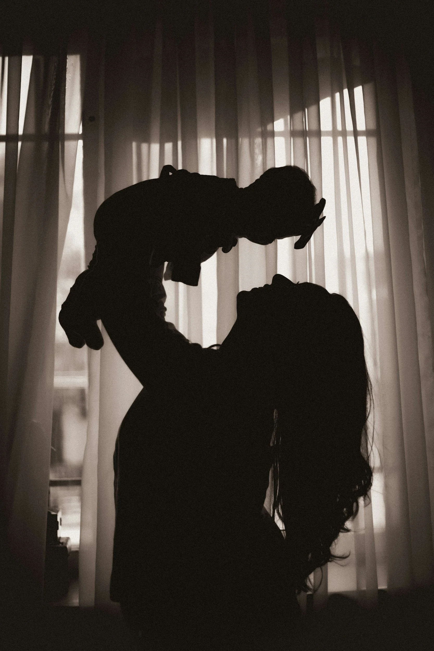Silhouette of a woman holding a child in front of sheer curtains, backlit by natural light.