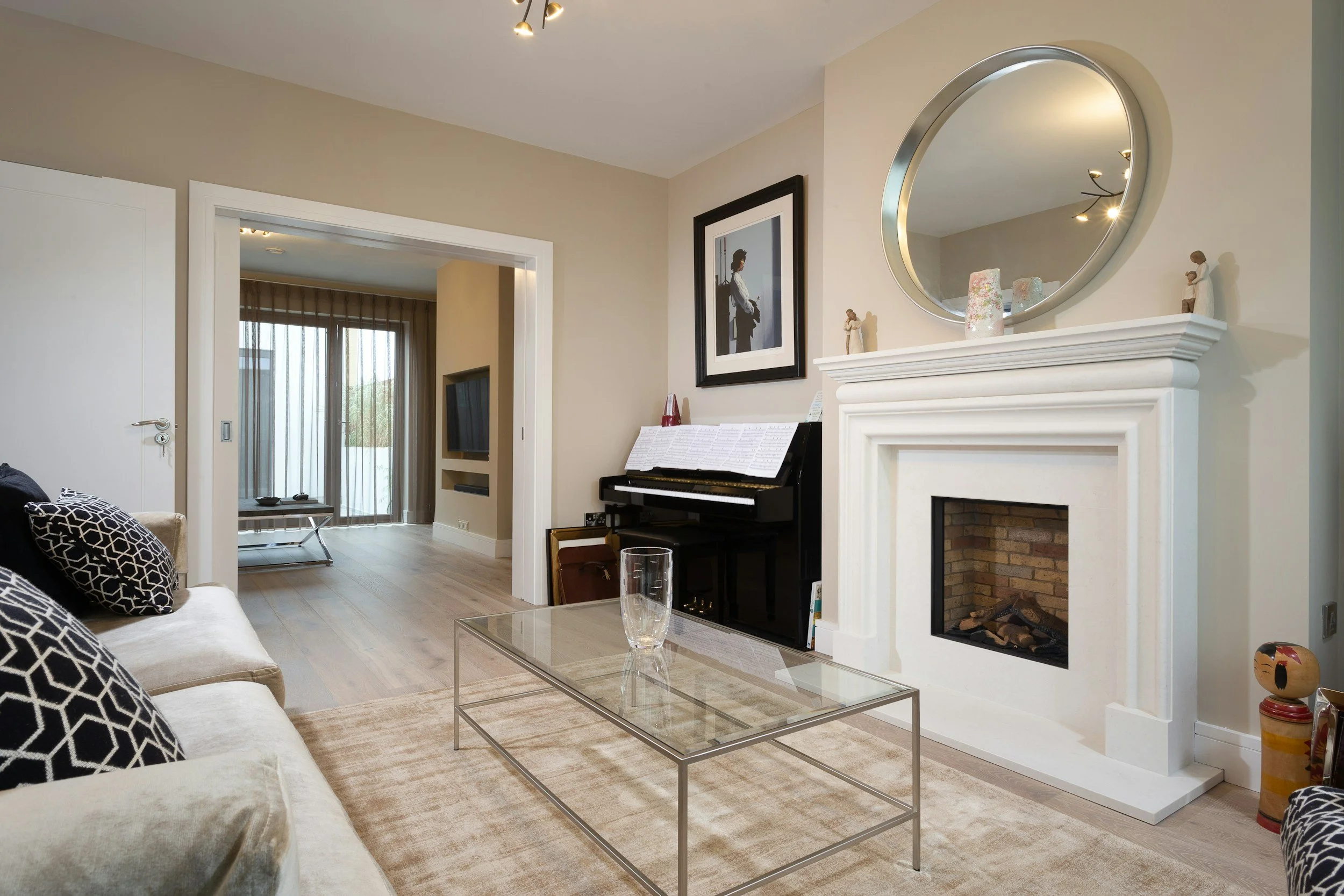 Living room with a white sofa, black and white patterned pillows, a glass coffee table, a fireplace, a round mirror above the fireplace, a piano with sheet music, a framed picture on the wall, and a doorway leading to a room with a TV and sliding glass doors.