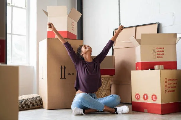 A woman sitting on the floor surrounded by moving boxes, celebrating with arms raised in a bright room.