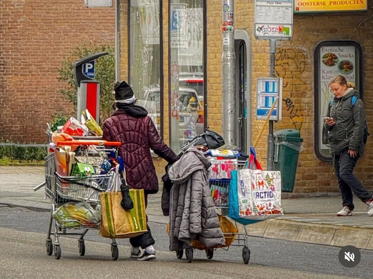 Twee vrouwen met boodschappentrolleys staan op de stoep bij een winkel, één kijkt op haar telefoon.