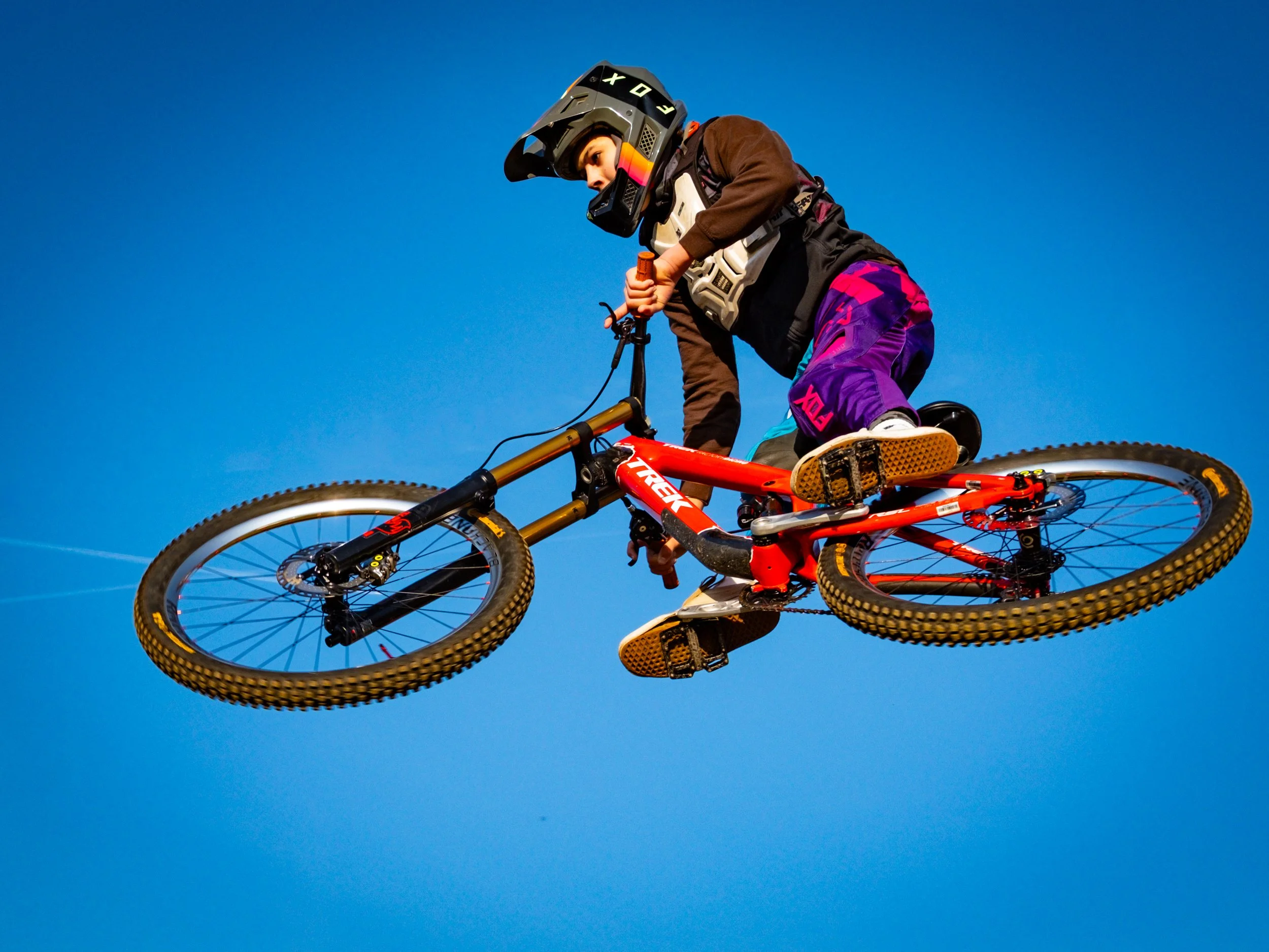 Kind op een mountainbike in de lucht tijdens een sprong met een heldere blauwe lucht op de achtergrond.