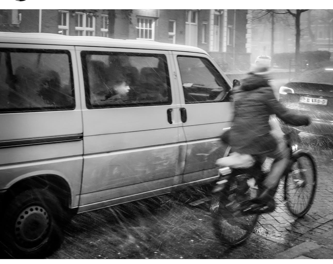 Een fietser rijdt snel langs een bestelauto op een besneeuwde straat met mensen zichtbaar door de raam van de auto.