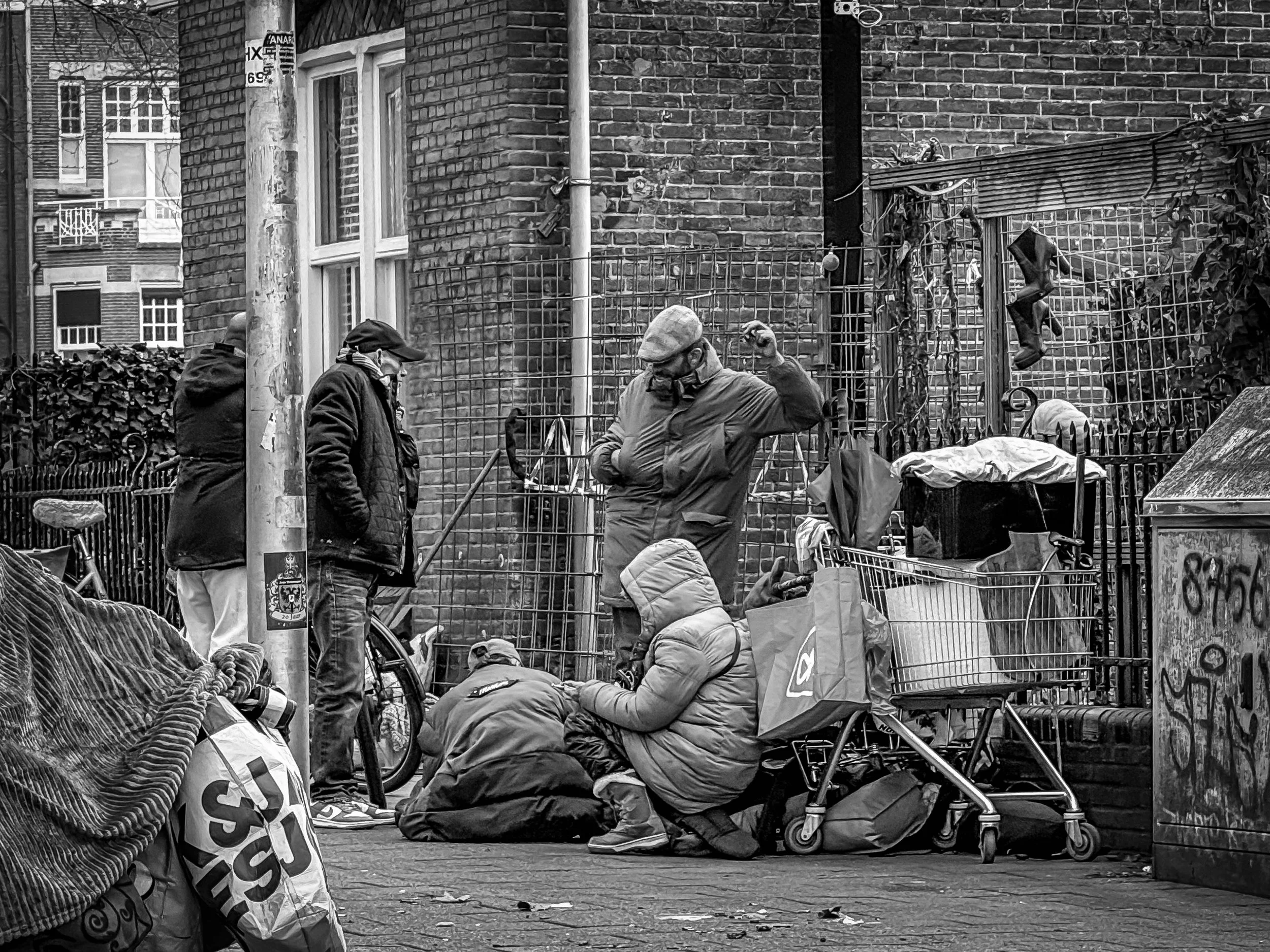 Vijf mensen op straat, twee staan en drie zitten, omringd door tassen, winkelwagens en een fiets, in een stedelijke omgeving met een bakstenen muur en een metalen hek op de achtergrond.