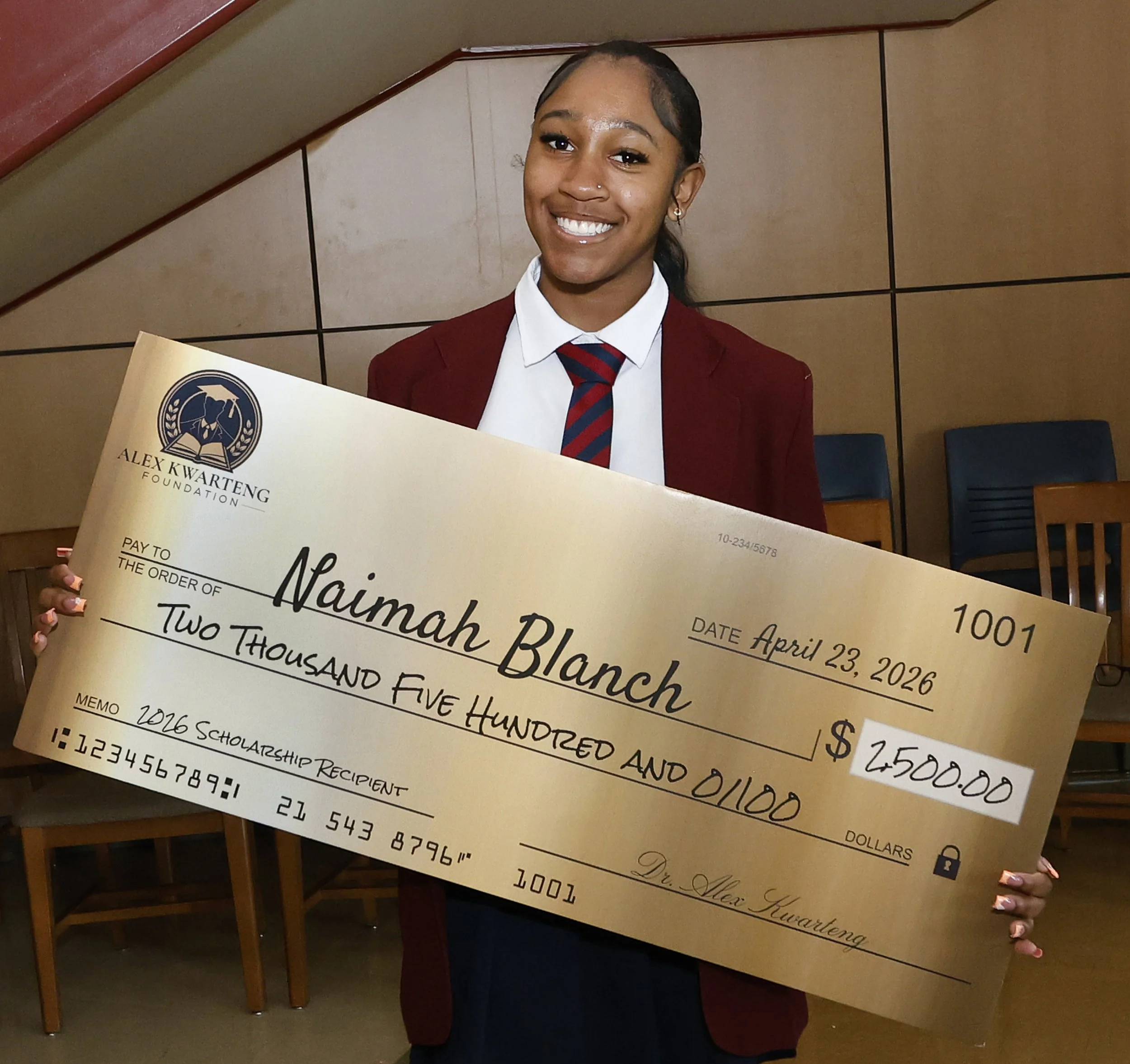 A woman dressed in a maroon blazer, white shirt, and red and navy striped tie holding a scholarship check made out to Naomi Blanch. She is smiling and has earrings and a small nose ring. The background is beige with wooden panels and chairs
