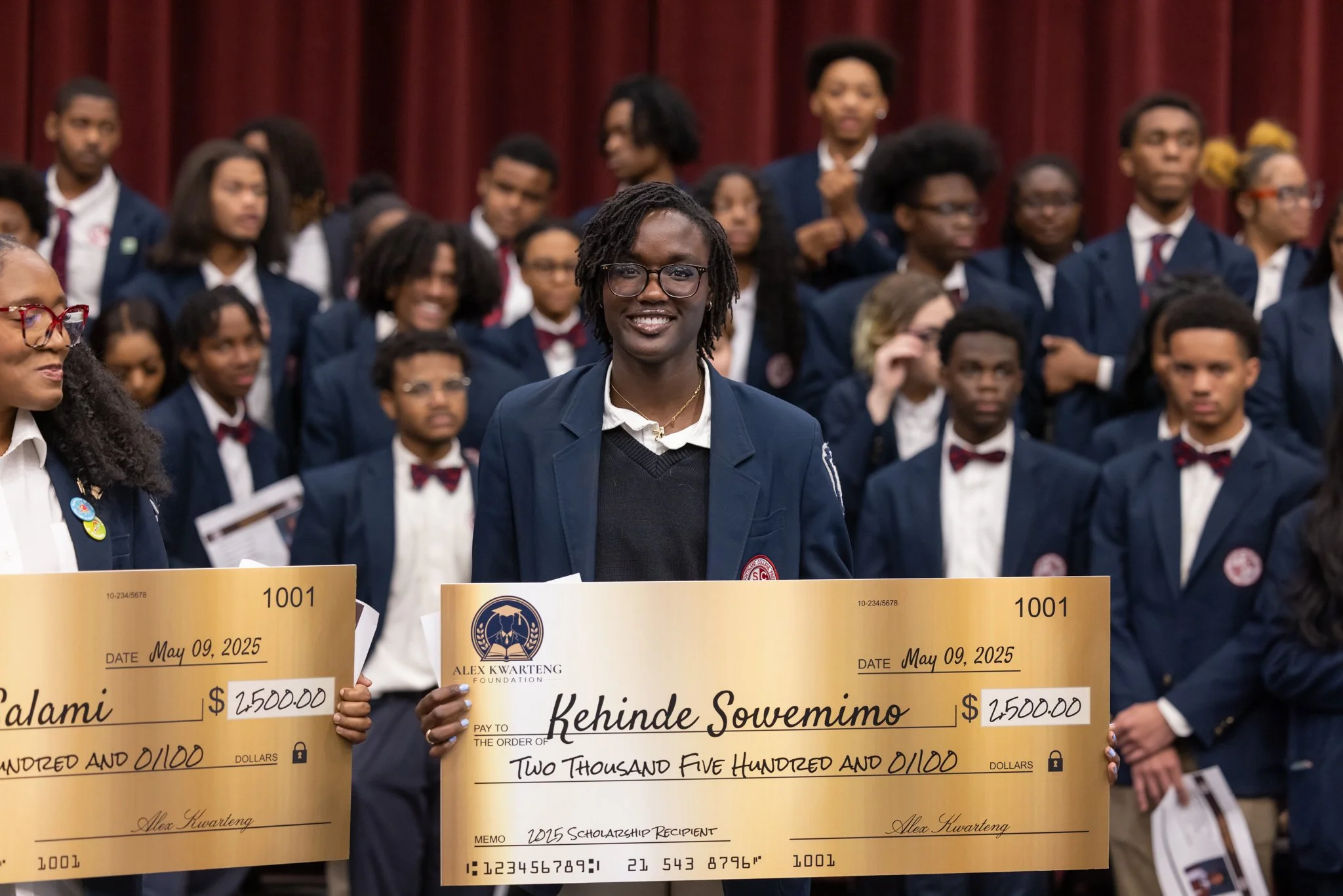 A young man with glasses and dreadlocks holds a large ceremonial check, smiling at an event with other young people in formal attire in the background.