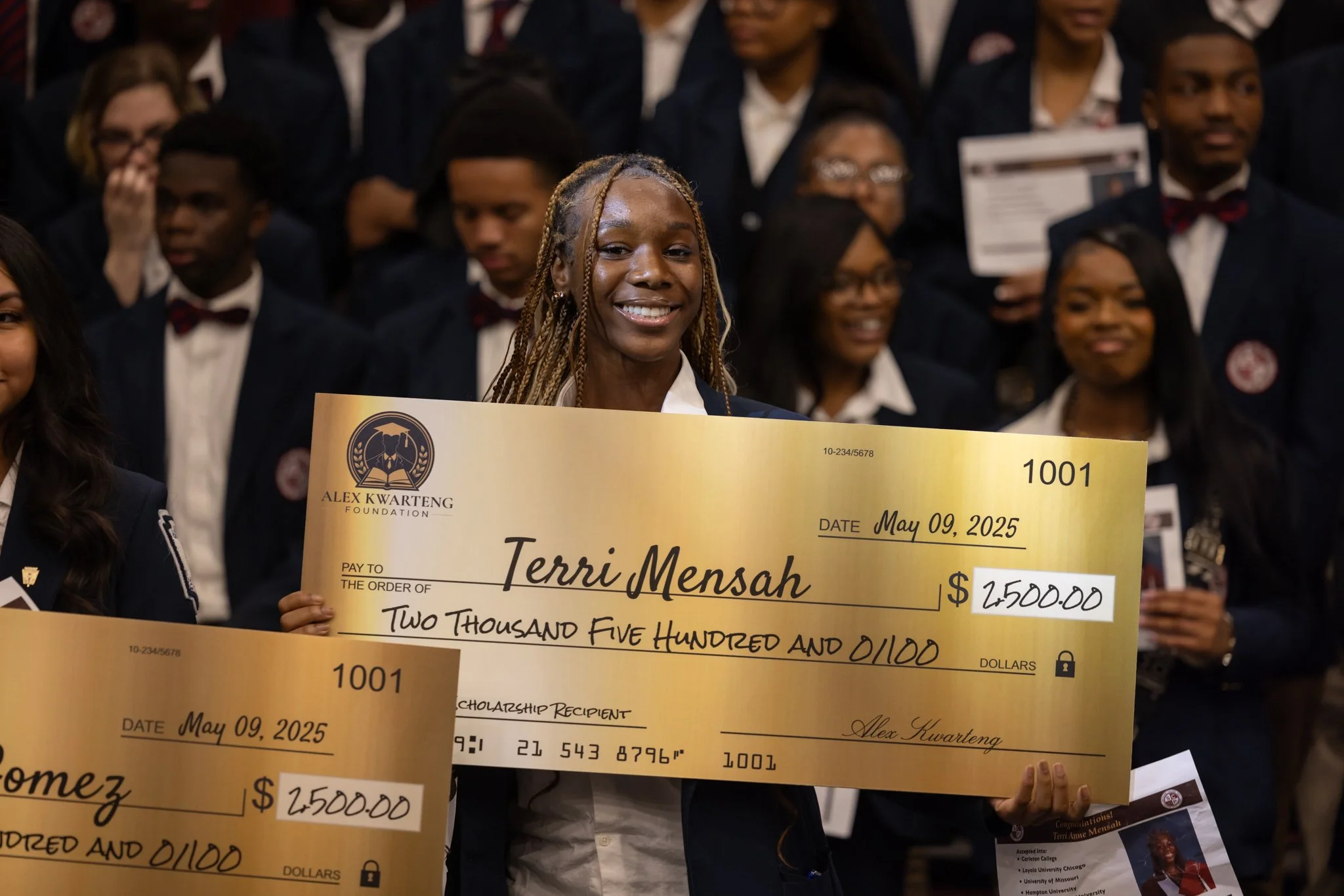 Young woman holding a large check at a graduation ceremony, surrounded by other graduates.