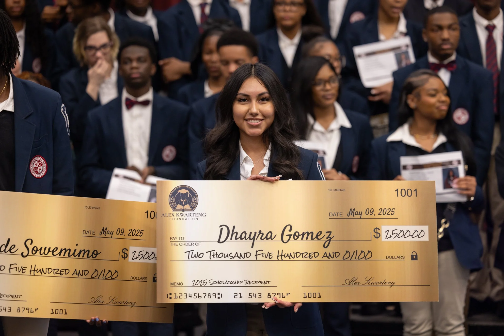 Young woman smiling, holding a large ceremonial check made out to Dhayra Gomez for $2,500, at a formal event with other people in suits in the background.