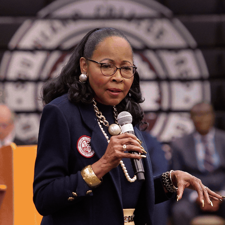 An African American woman wearing glasses, a navy blazer with a district patch, and large pearl jewelry, holding a microphone while speaking at a formal event with a logo in the background.