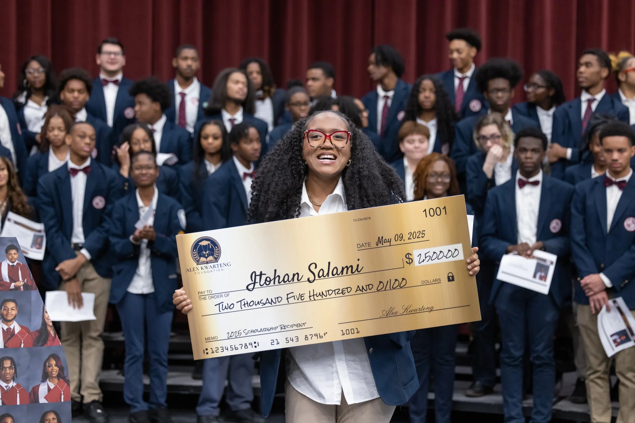 Smiling woman in glasses holding a large check for $2,500 at a formal event with many young people in suits and ties in the background.