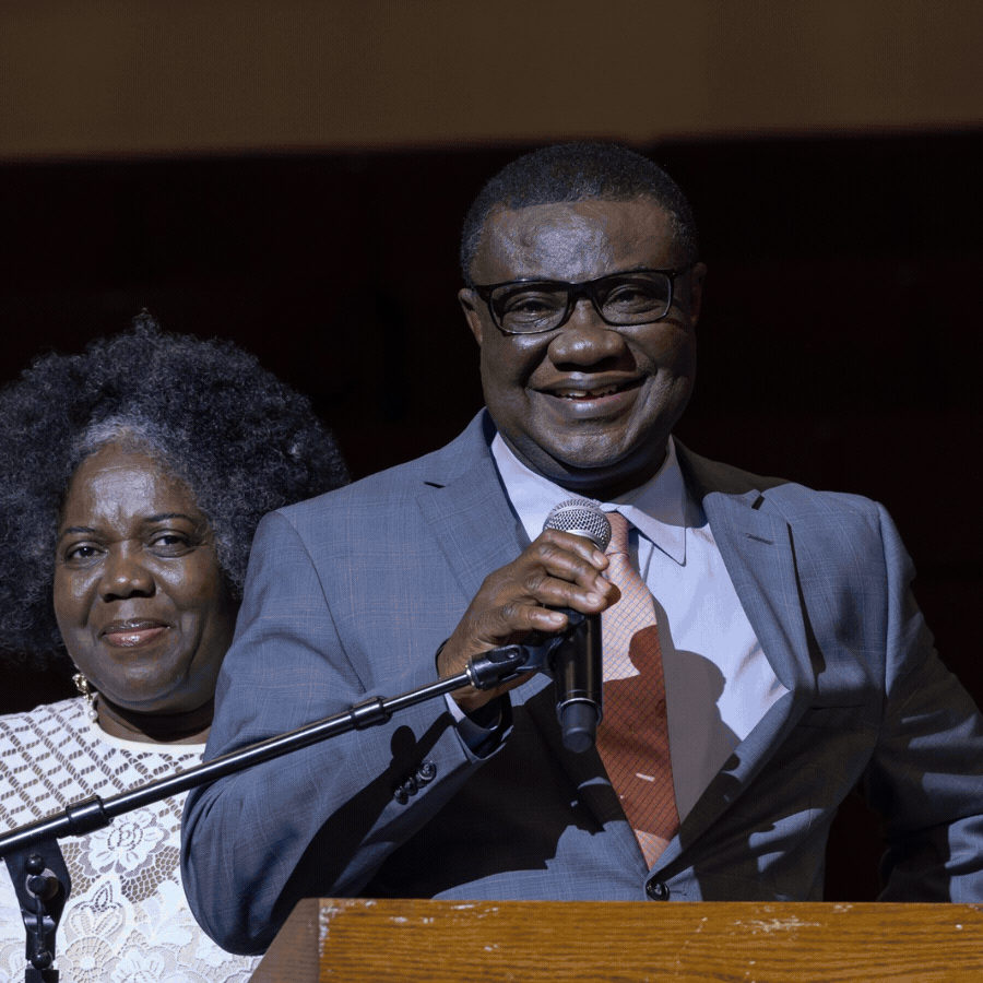 A man in a suit and glasses speaking into a microphone at a lectern, with a woman standing behind him, both smiling.