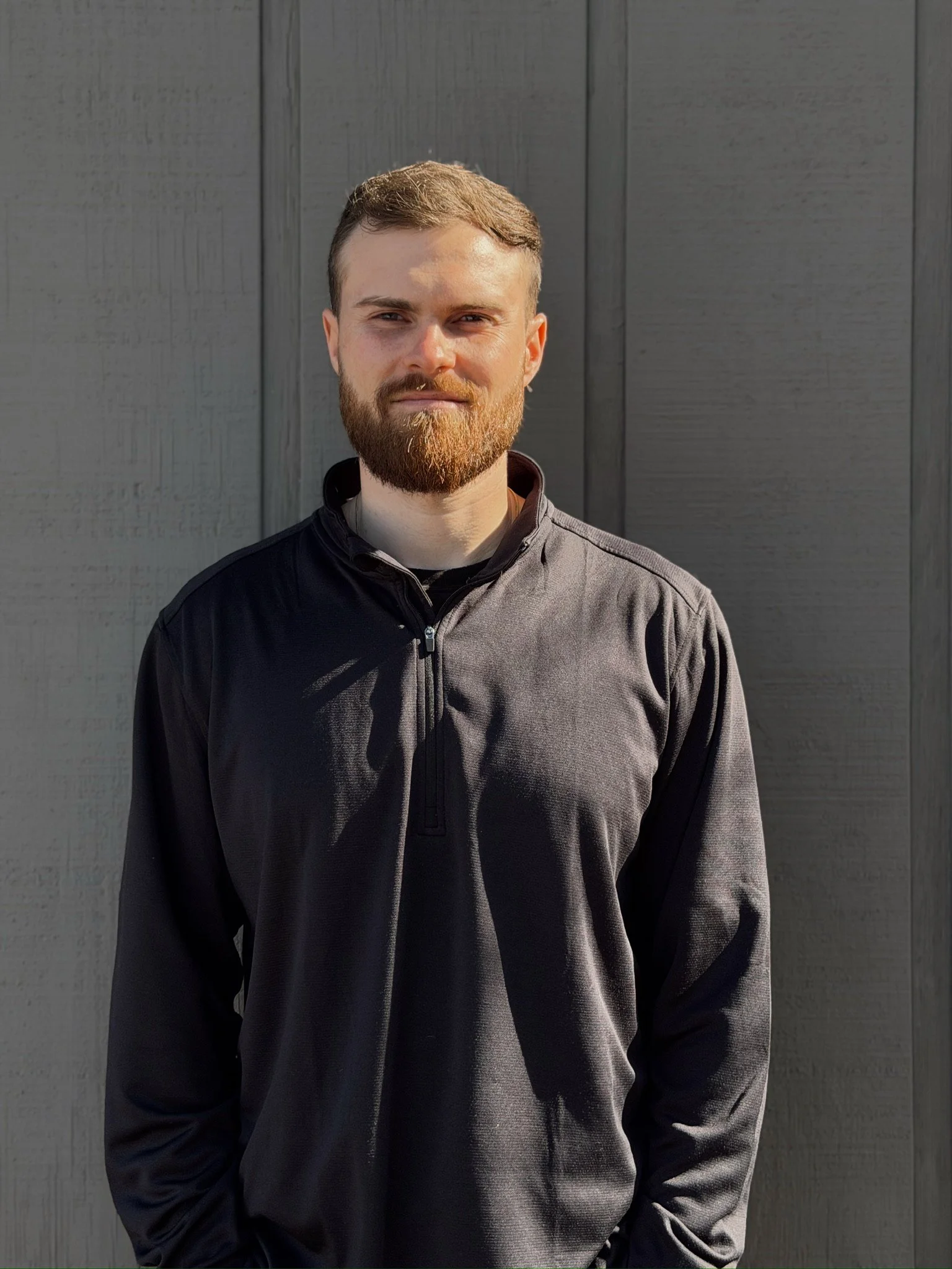 A man with a beard and short brown hair standing outdoors in front of a dark gray wooden wall, wearing a black quarter-zip jacket.