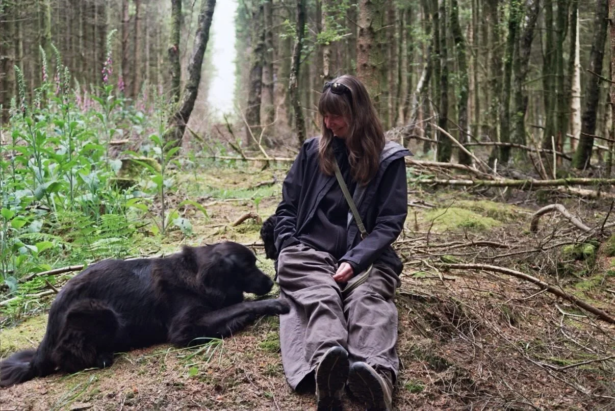 A woman in outdoor clothing sitting on the ground in a forest, playing with a black dog that is lying down. The scene features tall trees and green foliage.