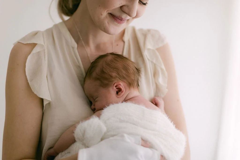 A woman gently holding a sleeping newborn baby on her chest, with soft lighting and a neutral background.