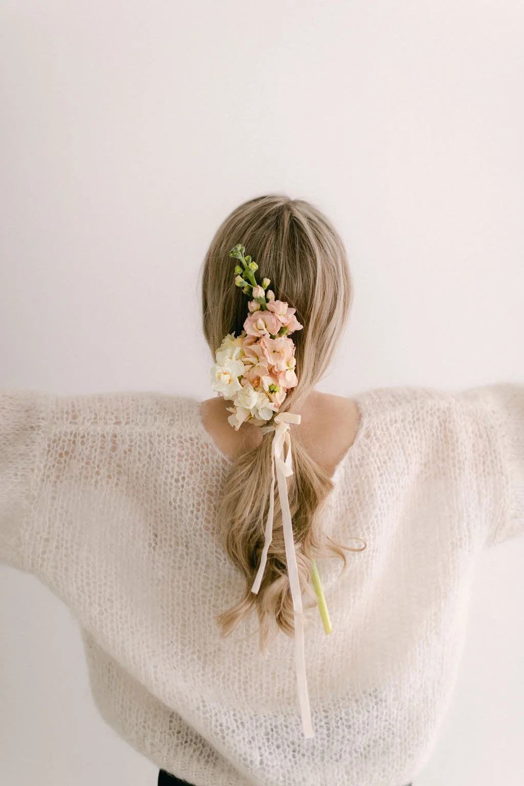 A woman with light blonde hair styled in a braid, decorated with pink and white flowers, tied with a white ribbon, wearing a light knit sweater, standing against a plain white wall.