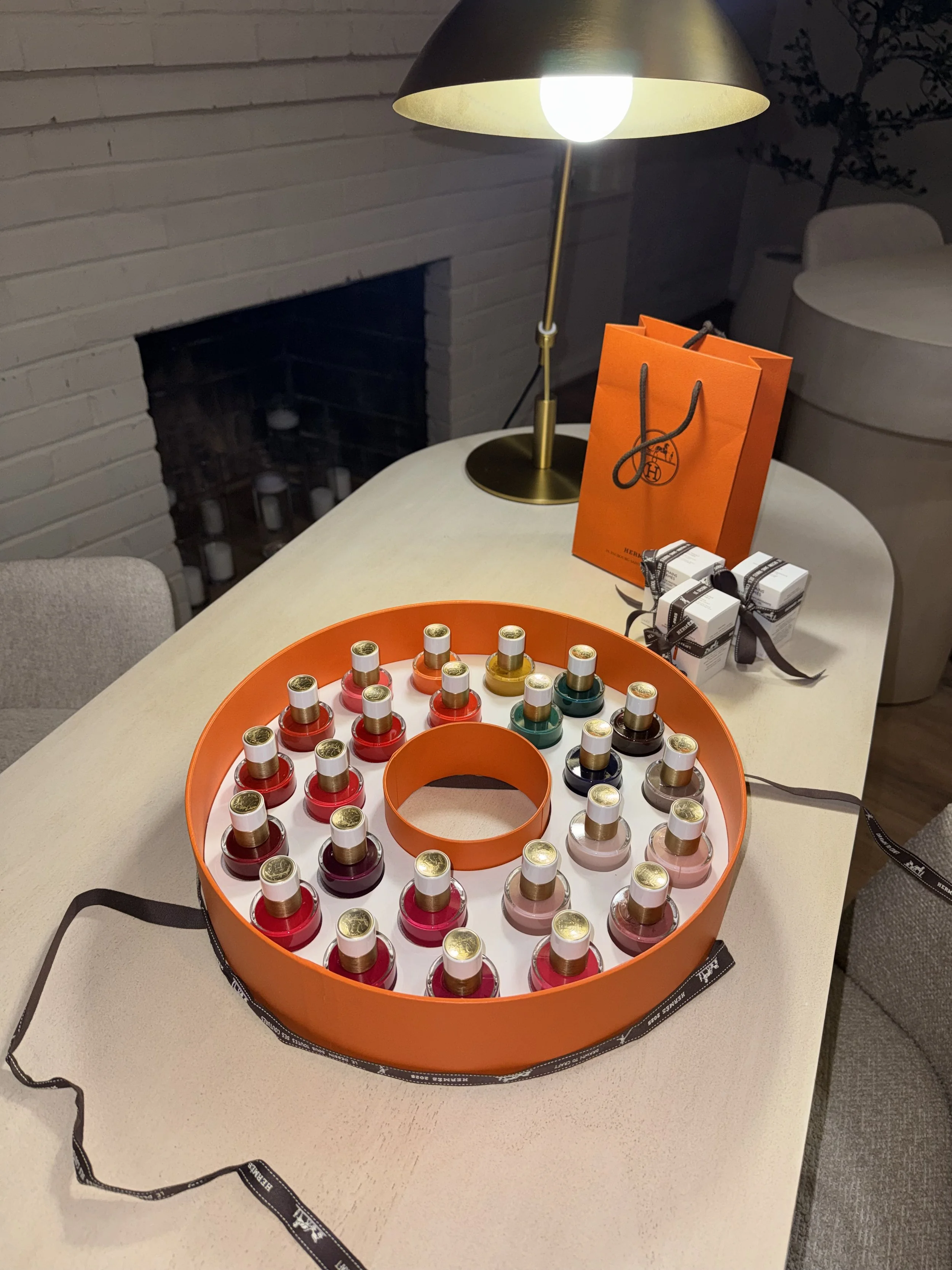 Display of multiple colorful bottles of nail polish arranged in a large orange circular box on a table, with a Hermes shopping bag and small wrapped gifts nearby, illuminated by a modern lamp.
