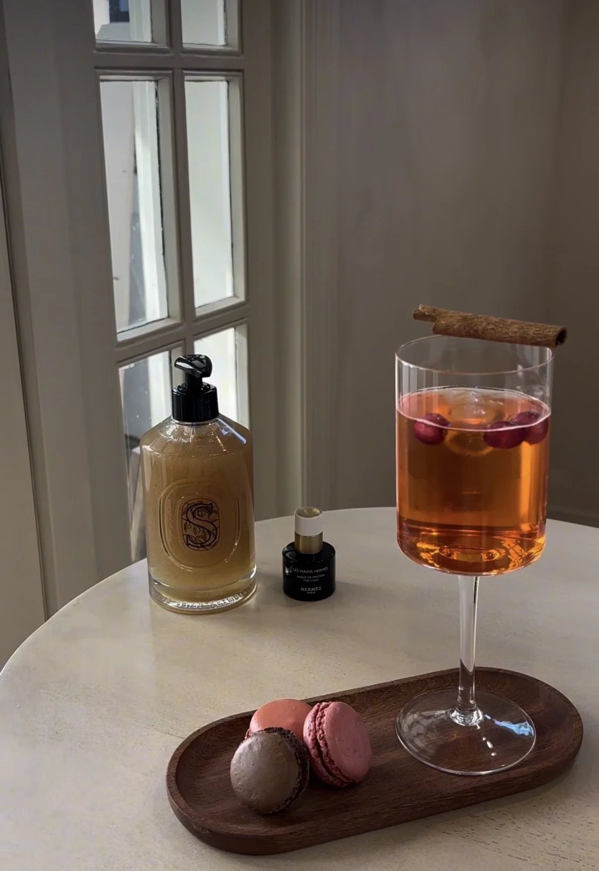 A table with a cocktail glass containing a pink-colored drink garnished with cranberries and a cinnamon stick, three macarons on a wooden tray, a large pump soap bottle, and a small bottle of perfume, with a window in the background.